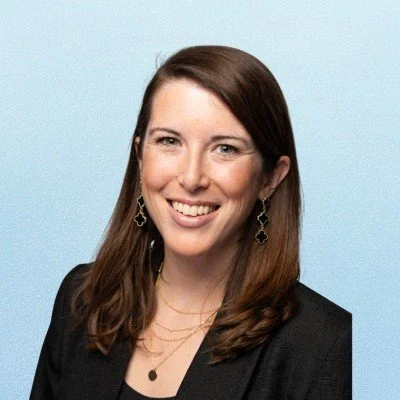 A woman with shoulder-length brown hair, smiling, wearing a black blazer, gold earrings, and layered necklaces, against a light blue background.