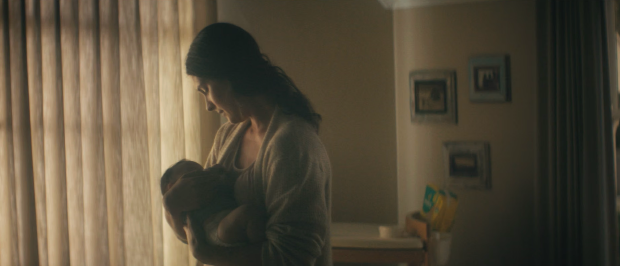 A woman holding a baby near a window with curtains.