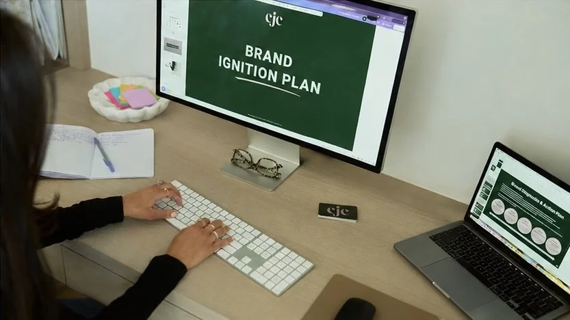 Person working at a desk with a desktop computer displaying a presentation titled 'BRAND IGNITION PLAN,' an open laptop showing a slide with circular diagrams, a notebook with a pen, glasses, a small black object with a logo, and a pet bed with stuffed animals in the background.