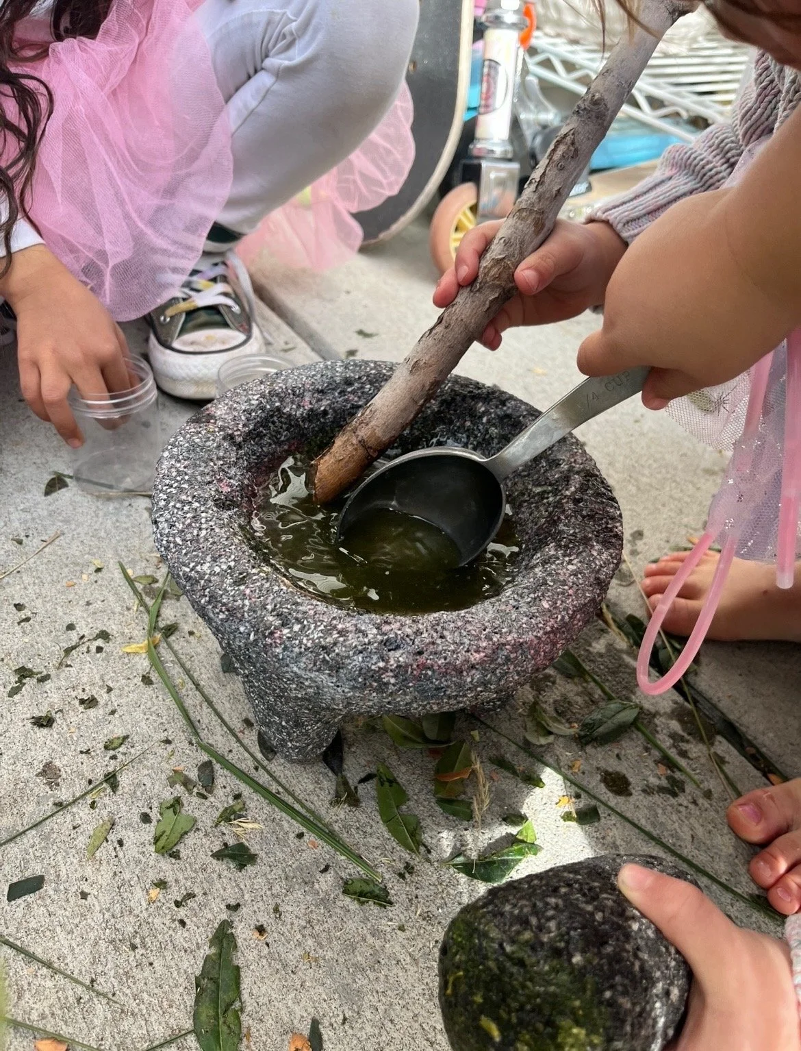 Children gathered around a stone mortar, crushing herbs with a pestle on a concrete surface scattered with leaves.
