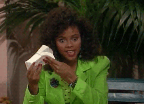 A woman with curly hair wearing a bright green blazer holding a book and gesturing with her finger, standing in front of large green plants.