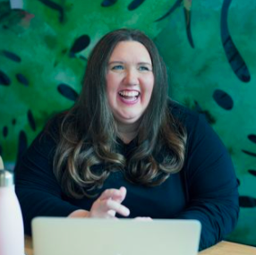A woman with long brown hair smiling and sitting at a table with a laptop in front of her, background features green tropical leaves.