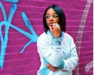 A woman with shoulder-length dark hair and glasses, wearing a light blue hoodie, stands in front of a pink brick wall with blue graffiti art.
