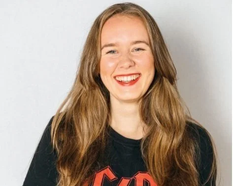 A young woman with long, wavy red hair, smiling, wearing a black T-shirt with red lettering.