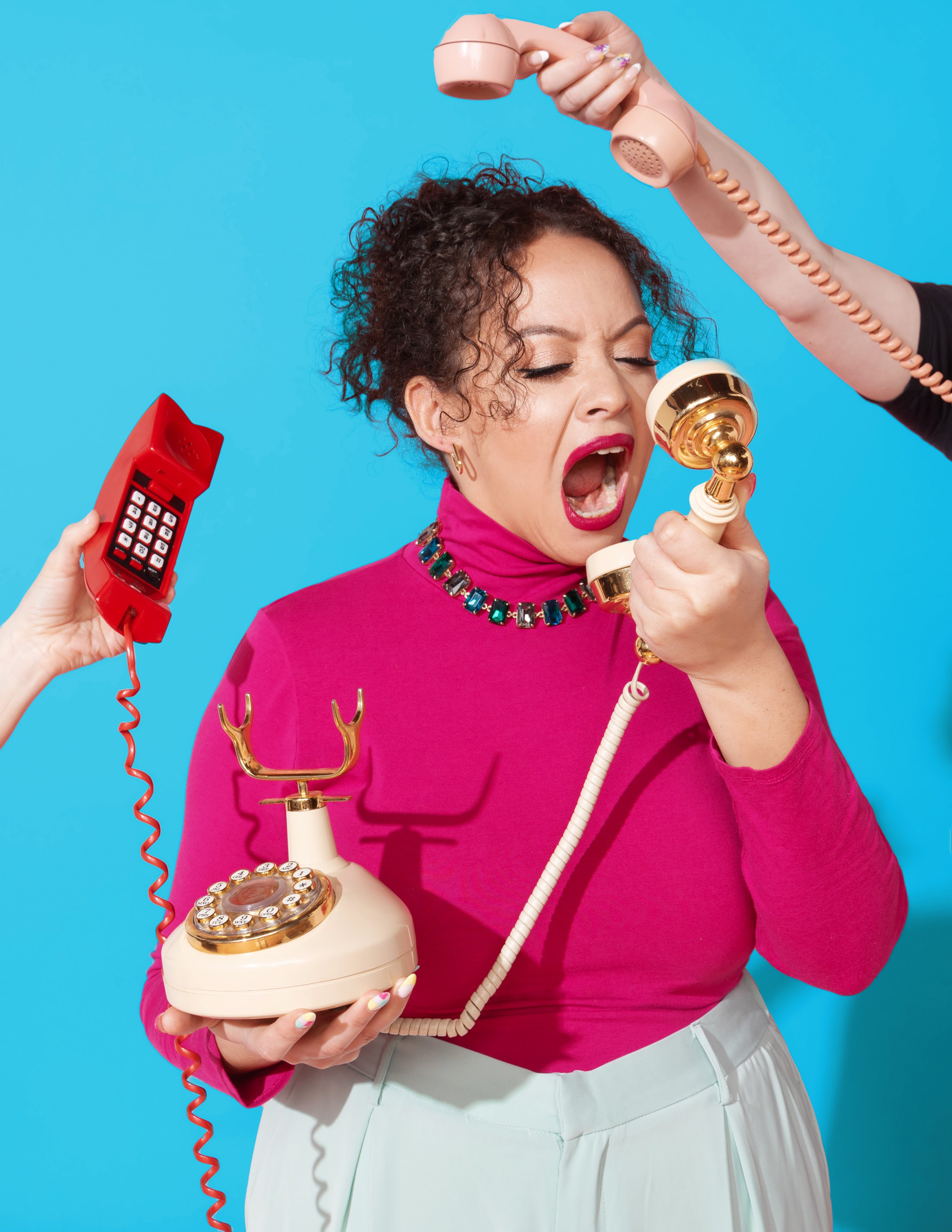 A woman wearing a pink top and white pants is shouting into a vintage gold and ivory rotary phone, holding it with her left hand while a hand holding a red cordless phone is raised near her face and another hand holding a pink telephone handset is placed above her head, against a bright blue background.