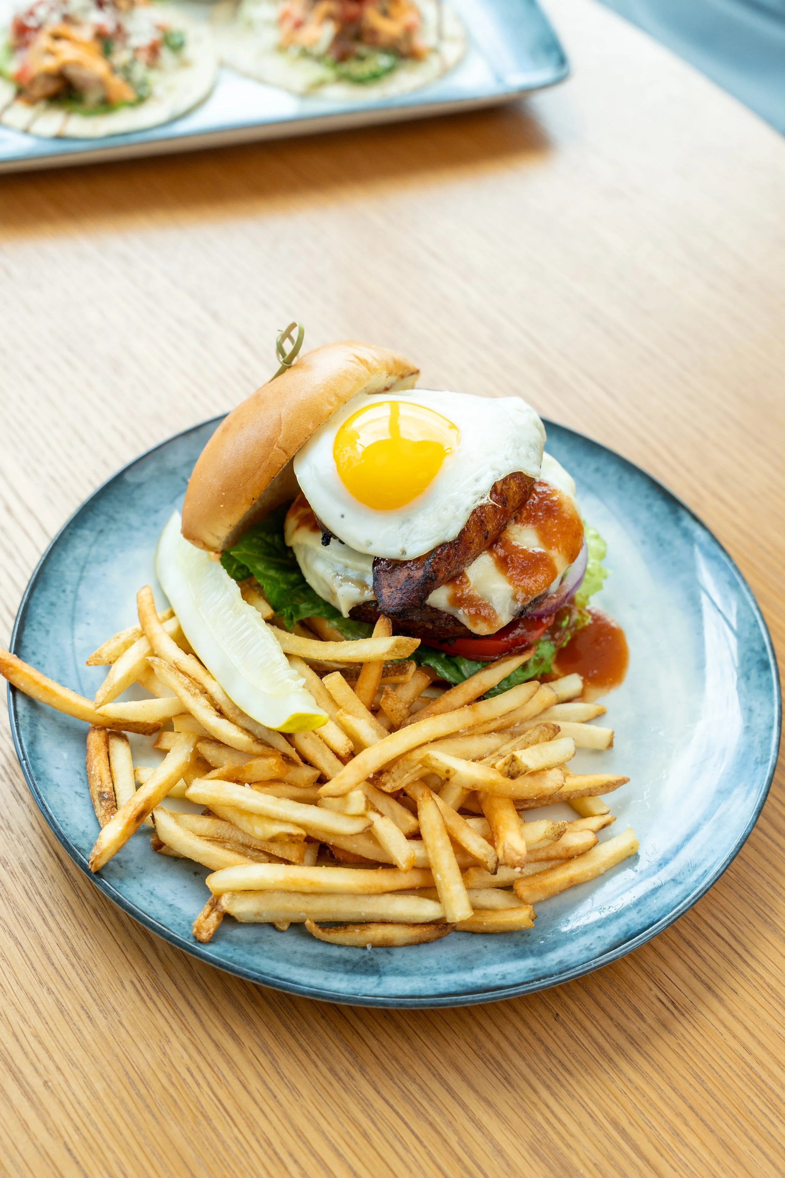 Florida Burger with a sunny-side-up egg on top, lettuce, tomato, cheese, and bacon, served with French fries and pickles on a blue plate.