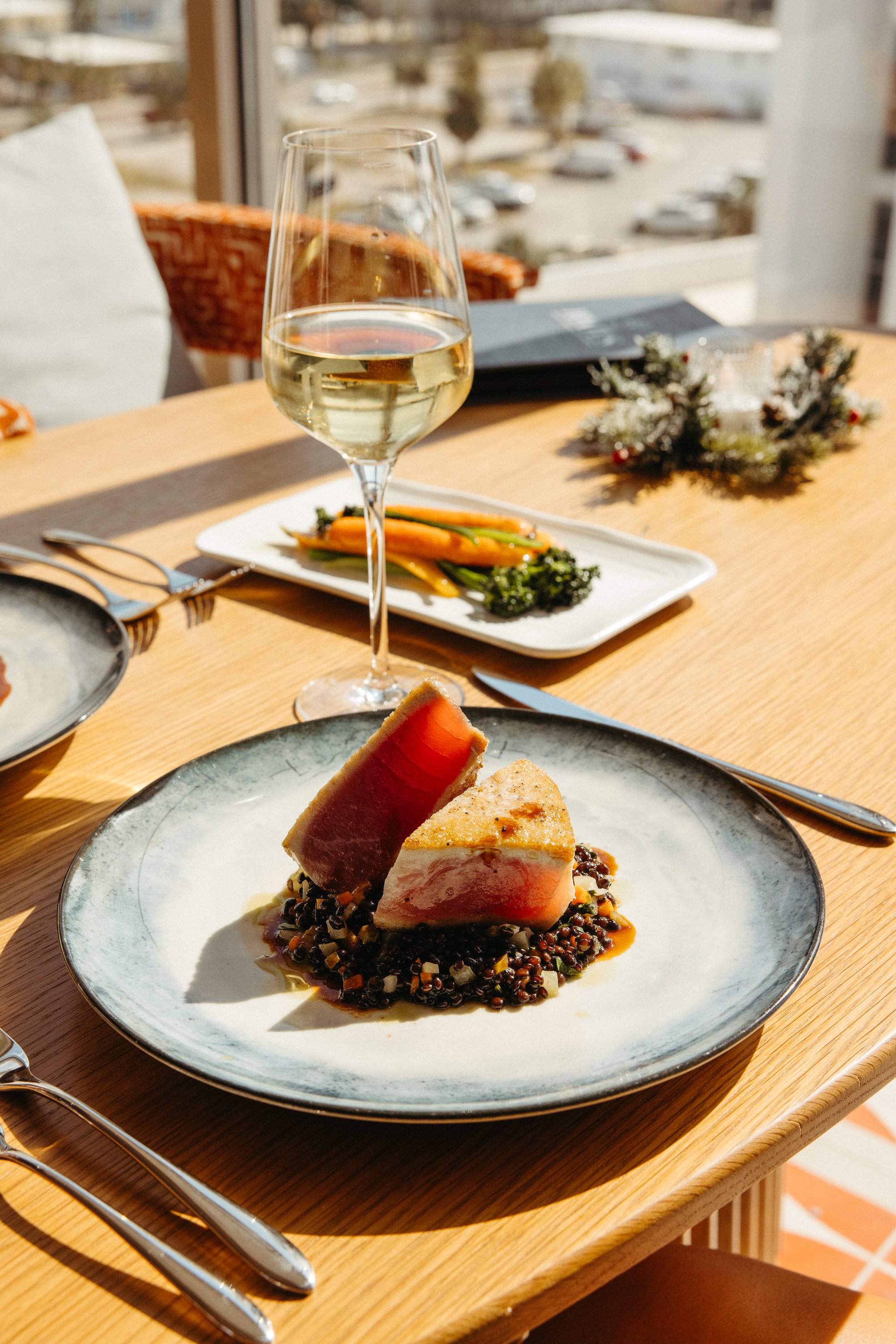 A plated dish with two pieces of seared tuna on a bed of grains, alongside a glass of white wine on a wooden table with additional plates of vegetables and festive decorations.
