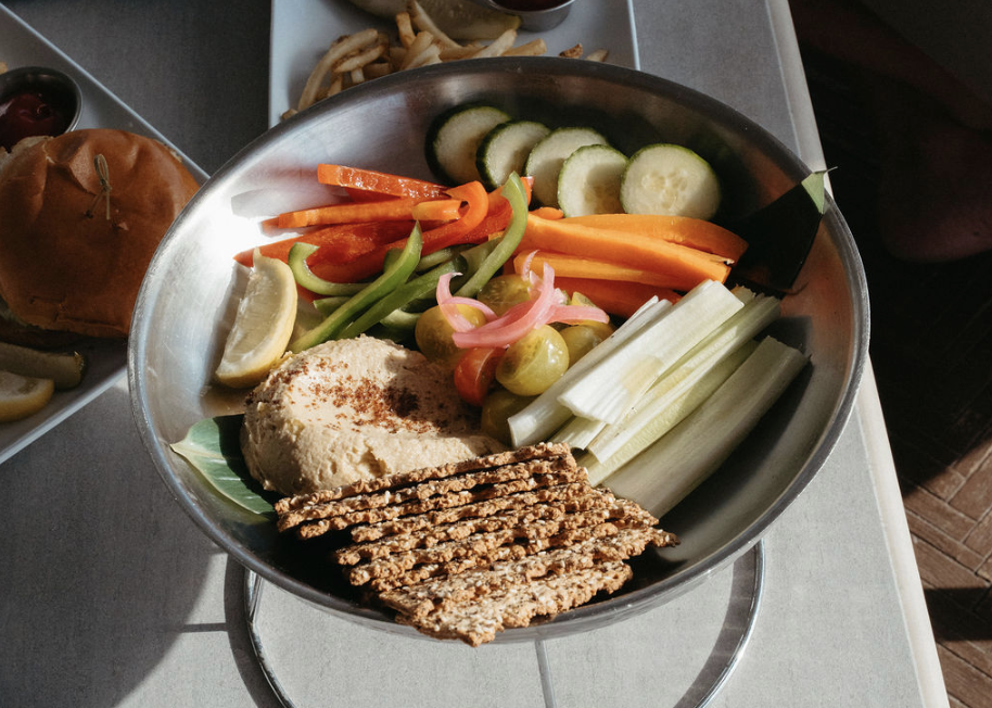 A platter of sliced cucumber, celery, carrots, pickled onions, and a scoop of hummus, served with whole grain crackers.