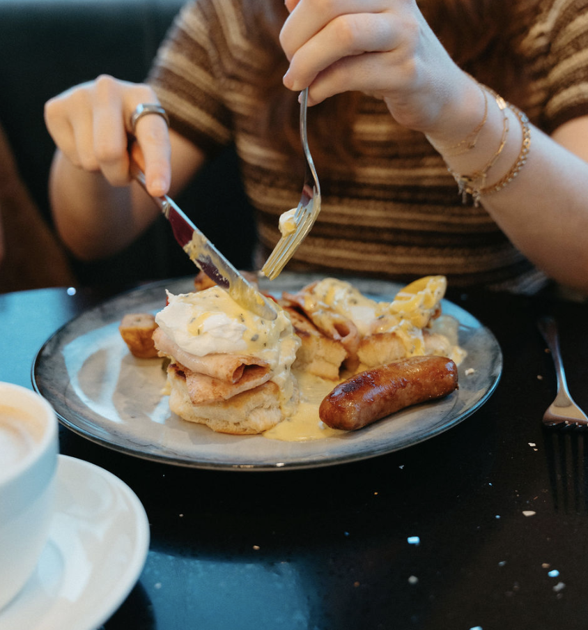 Person eating breakfast at Ocean Seven, lifting a fork and knife to a plate with eggs benedict, and sausage.