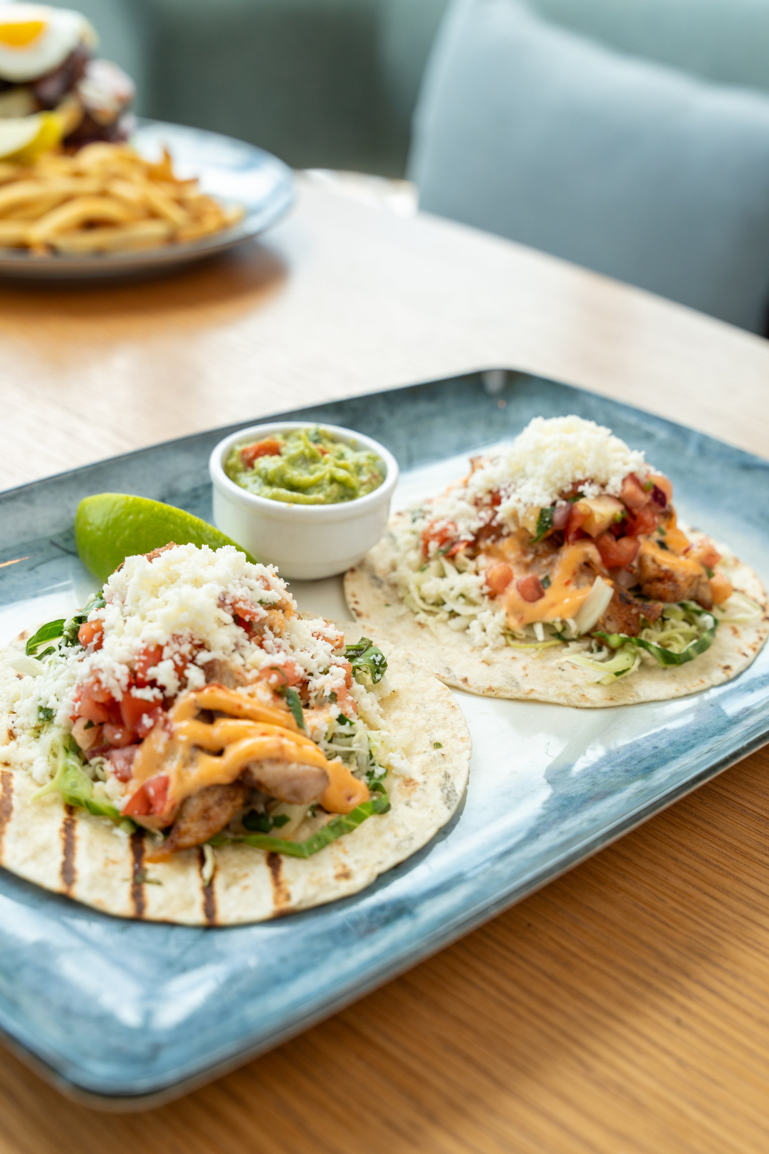 Two tacos with grilled Mahi Mahi, lettuce, tomatoes, shredded cheese, and drizzled sauce on corn tortillas, served with guacamole and lime on a rectangular blue plate.