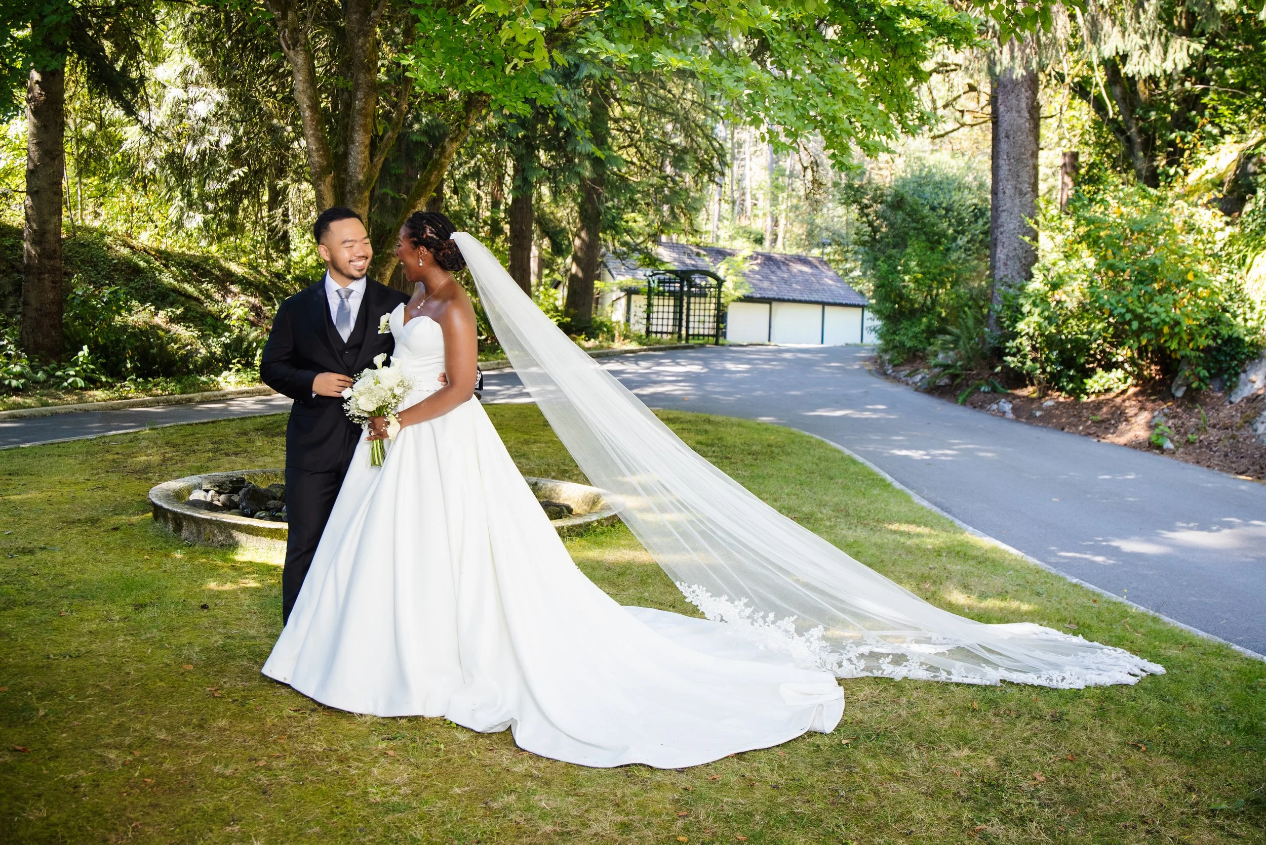 A newlywed couple standing outdoors in a lush green park, smiling at each other. The bride is wearing a white wedding dress with a long veil, holding a bouquet of white flowers. The groom is dressed in a black suit with a light-colored tie. They are 