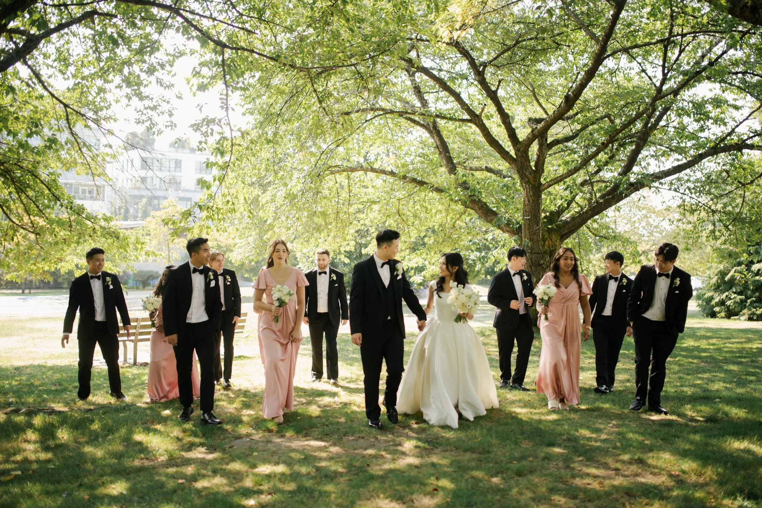 Bridal party dressed in formal wear walking under a large tree in a park on a sunny day. Wedding day coordination by Ona Events