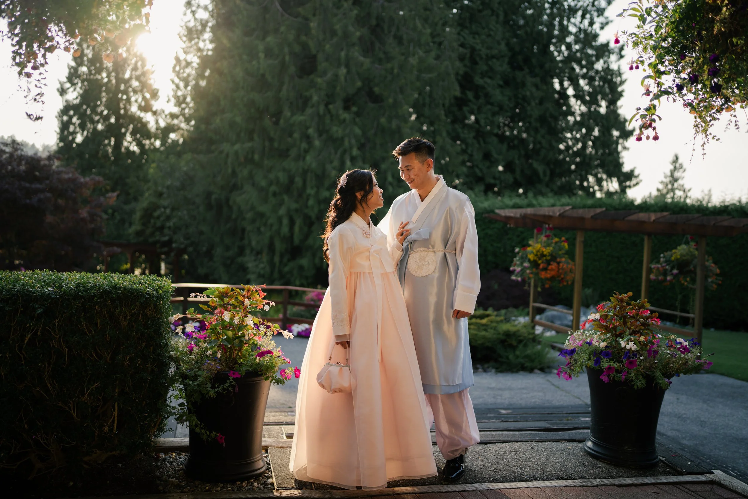 An Ona Events wedding couple dressed in traditional Korean hanbok, smiling and looking at each other in an outdoor garden setting during late afternoon.
