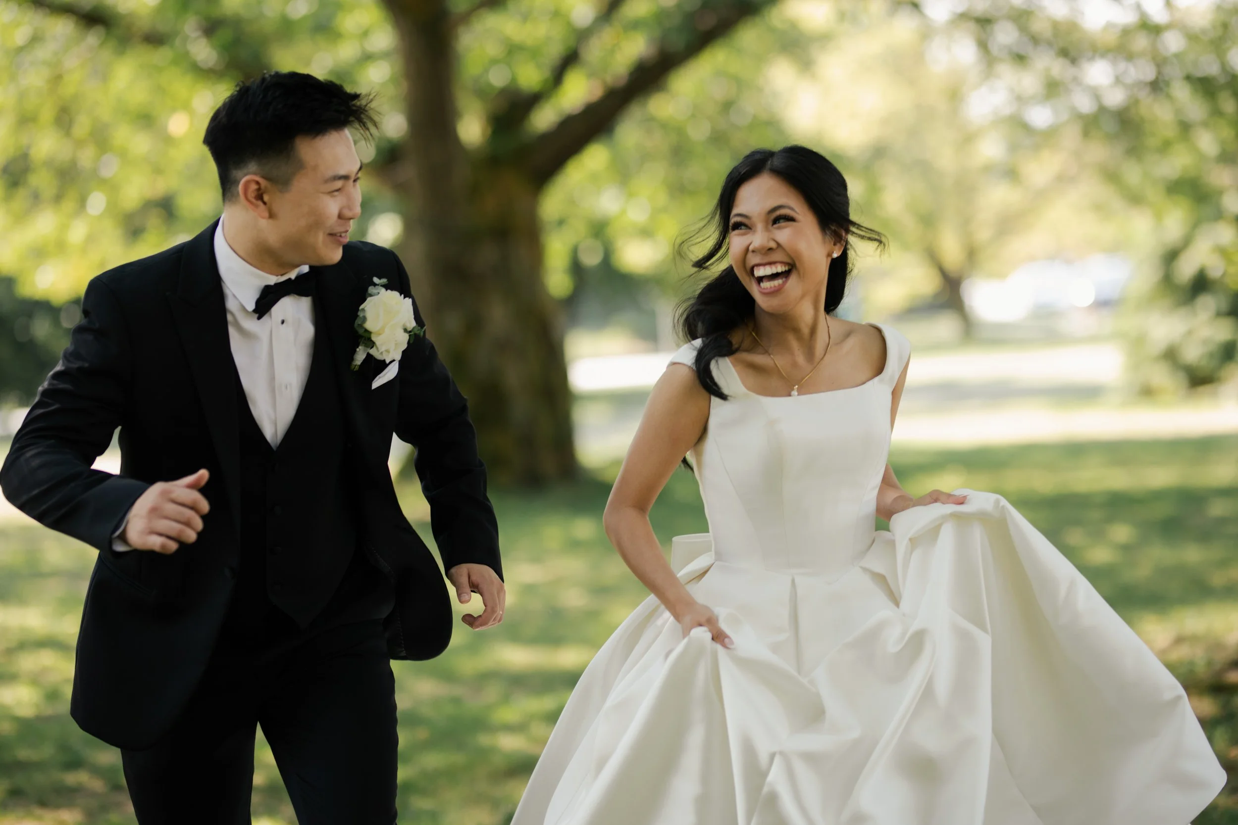 An Ona Events bride and groom in wedding attire are joyfully running outdoors amidst green trees, smiling at each other.