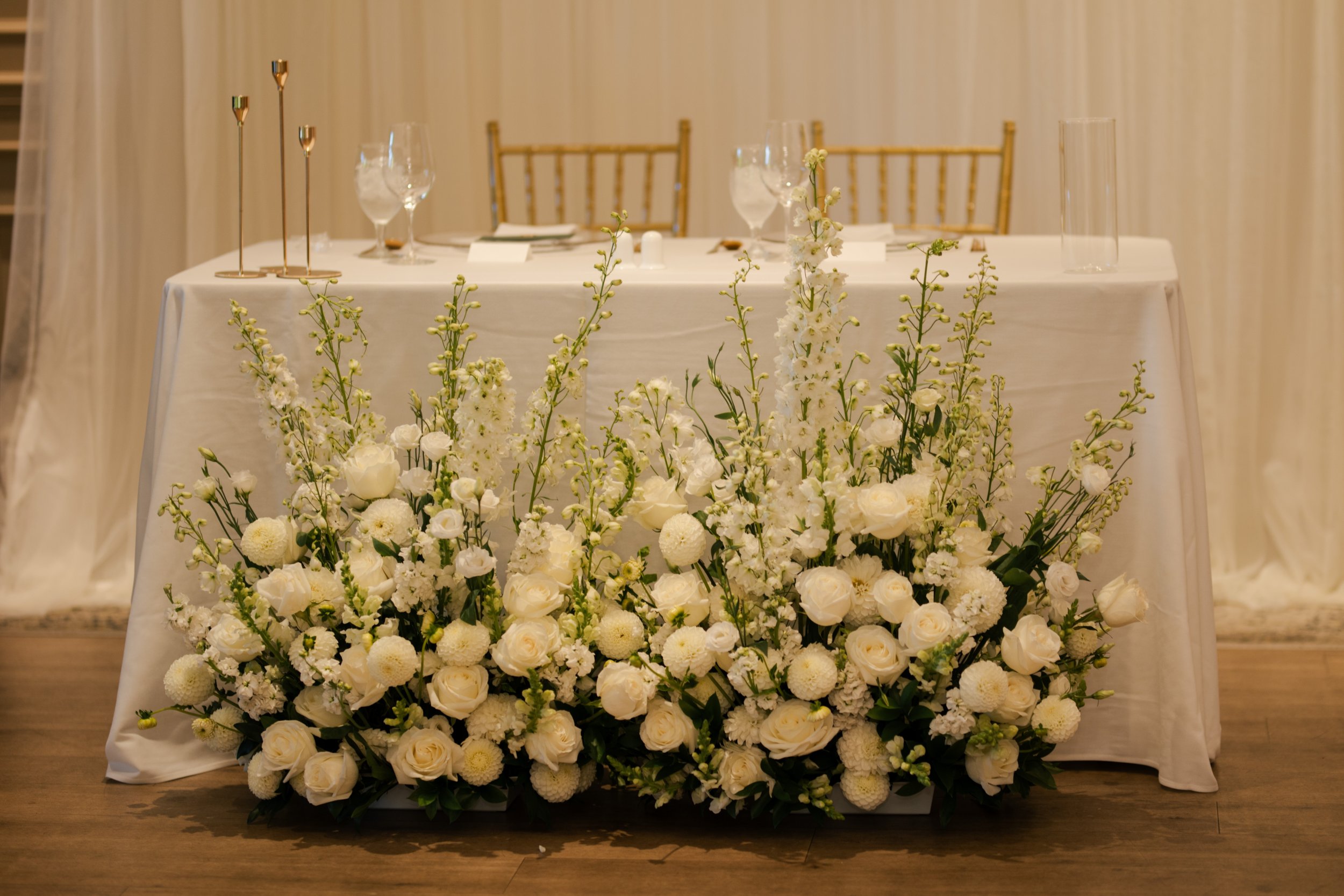 A long banquet table decorated with white flowers and white tablecloths, set for a formal event with wine glasses and gold accents.