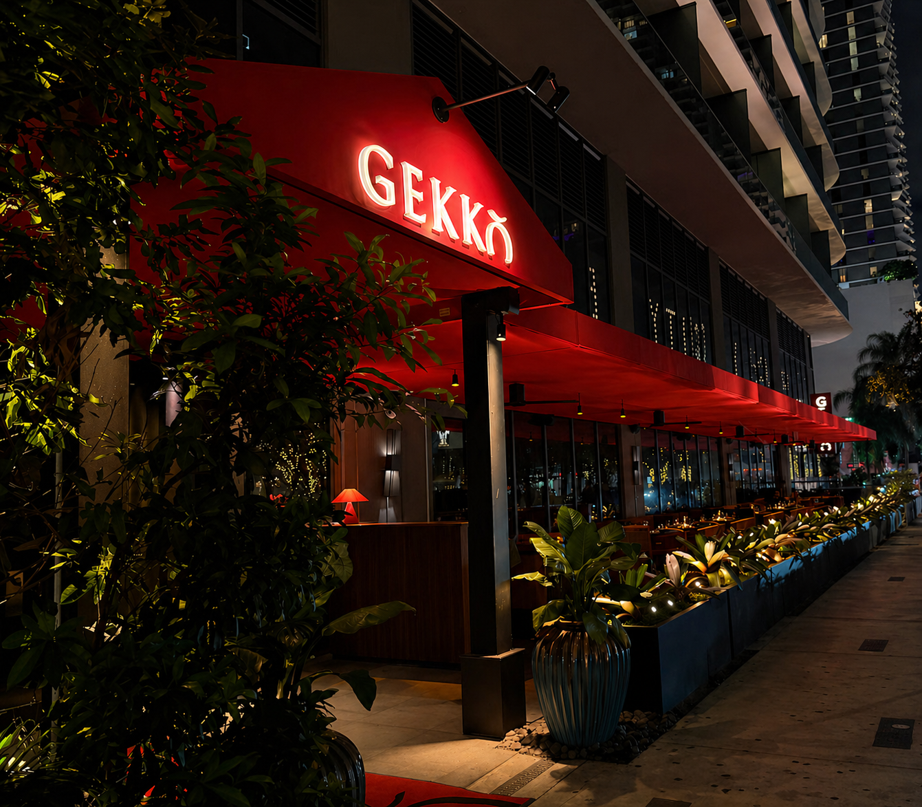 Nighttime view of a restaurant with a red awning and illuminated sign that reads 'GEKKO', with outdoor seating, plants, and city buildings in the background.