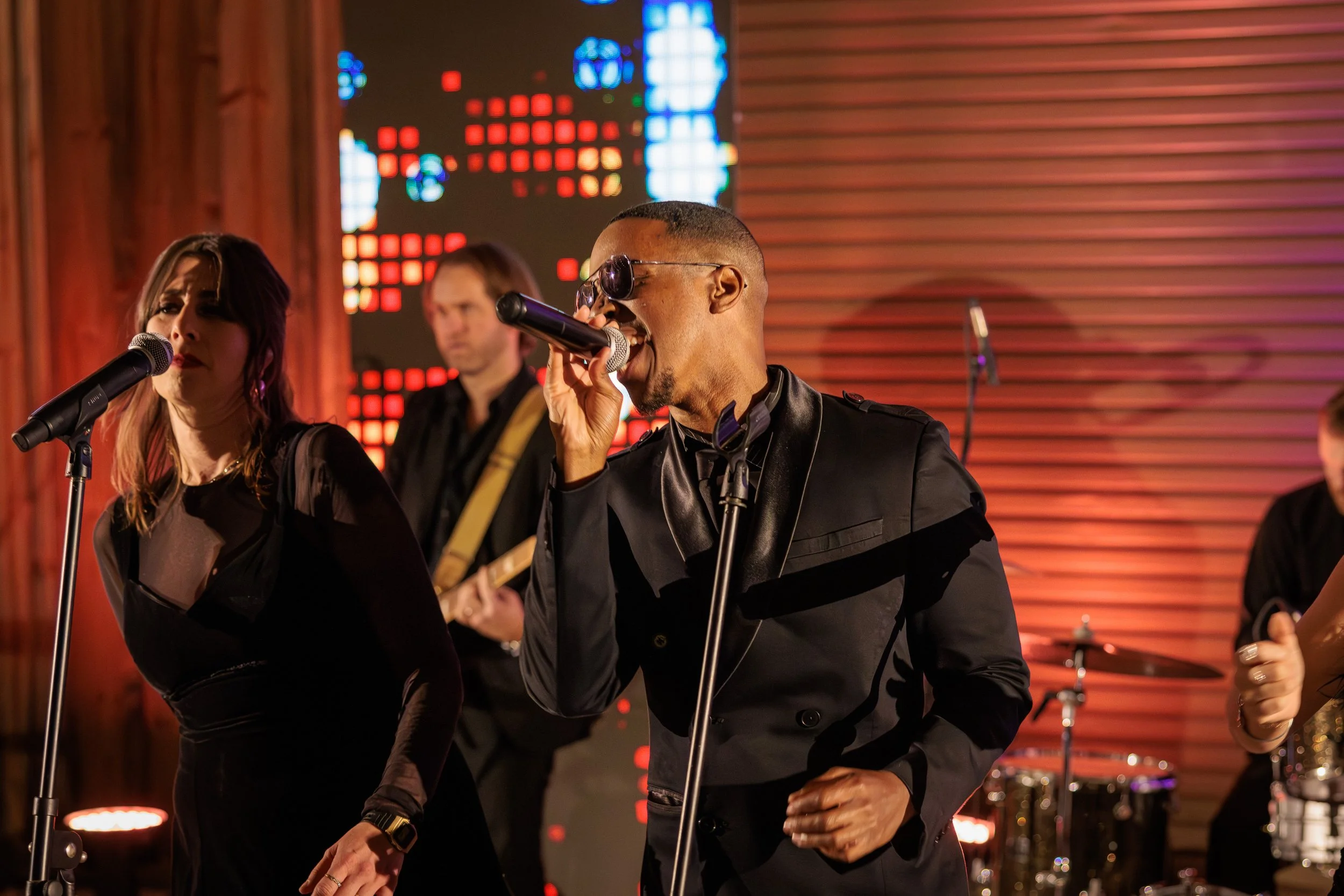 Musicians performing on stage, with a male singer wearing sunglasses, black jacket, and holding a microphone, female singer in black dress, and other band members playing instruments in the background, illuminated by stage lights.