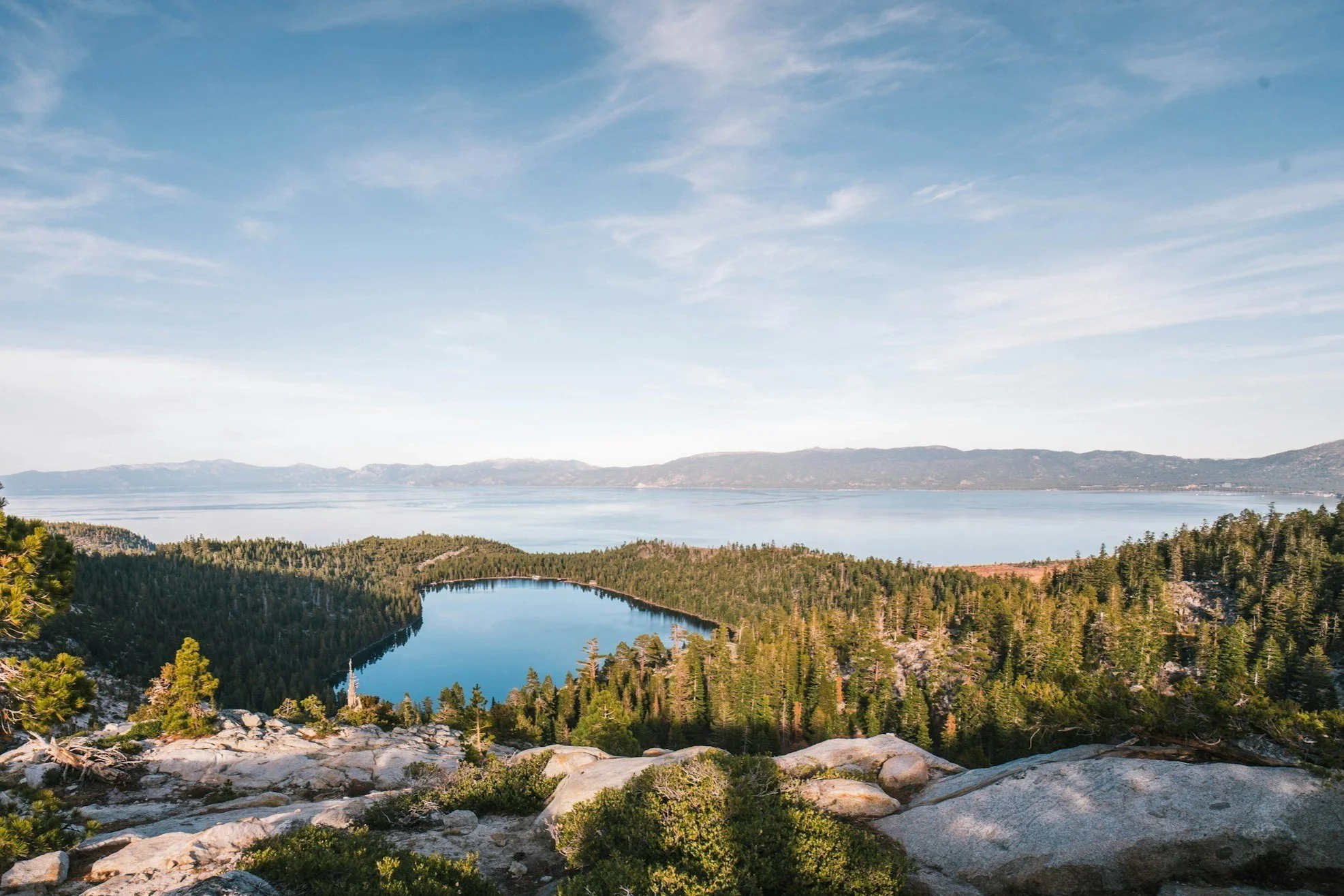 Scenic landscape of a lake surrounded by dense pine trees, with mountains in the distance under a blue sky with wispy clouds.