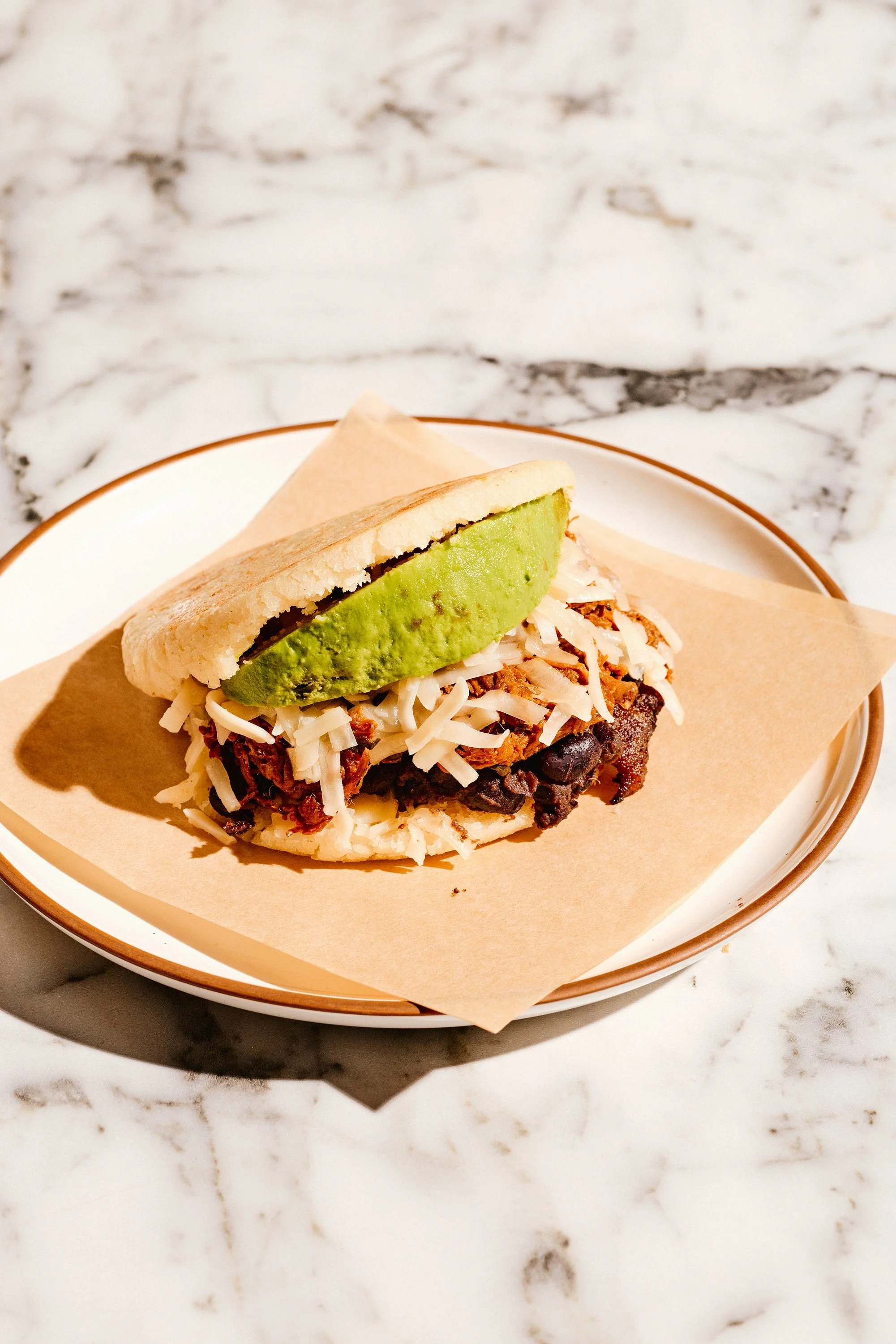 A taco with shredded cheese, pulled meat, and a slice of avocado on top, served on a paper-lined dish on a marble surface.