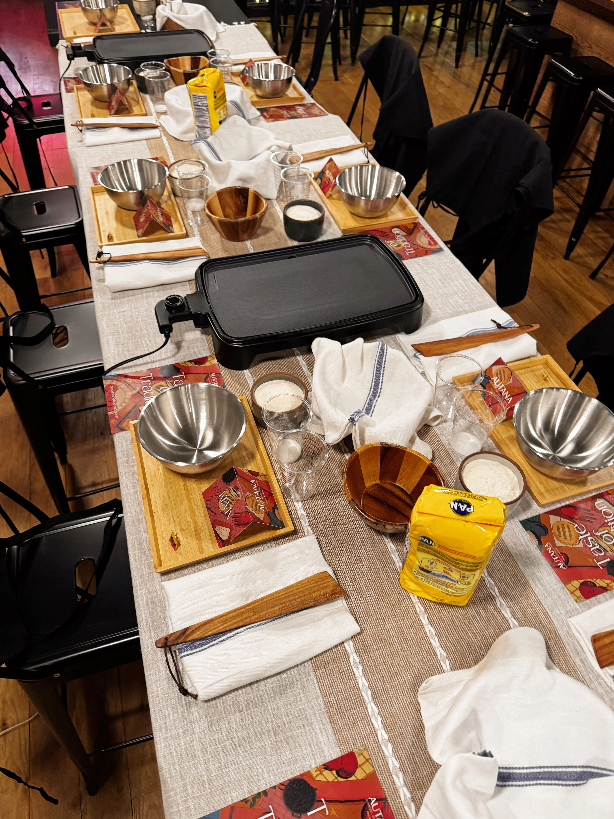 Arepa workshop. A dining table set for a meal with wooden boards, stainless steel bowls, glasses, napkins, and a black electric griddle in the center, in a restaurant with wooden floors and chairs.