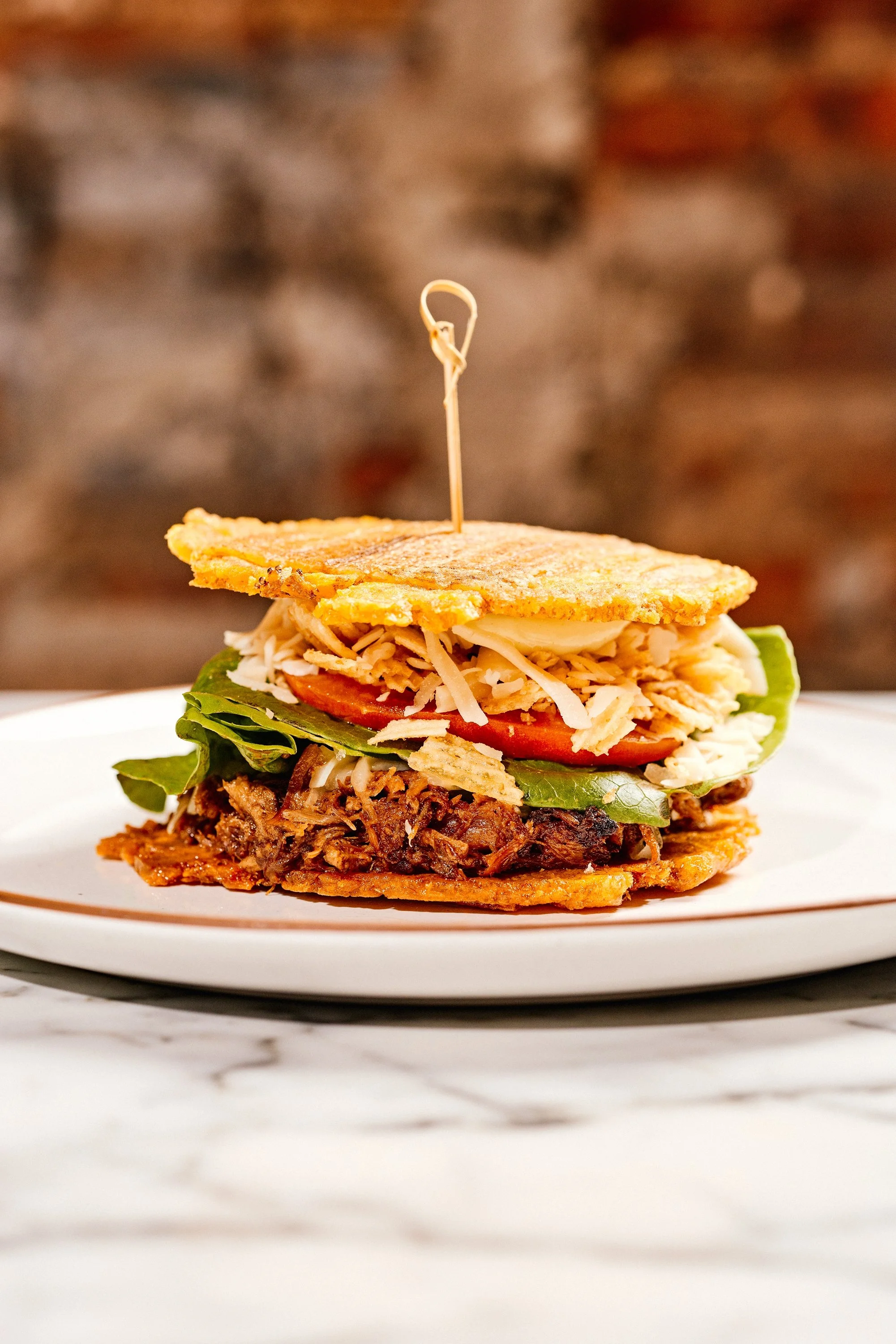 An arepa stuffed with shredded beef, lettuce, tomato, cheese, and fried plantains, held together with a toothpick on a plate.