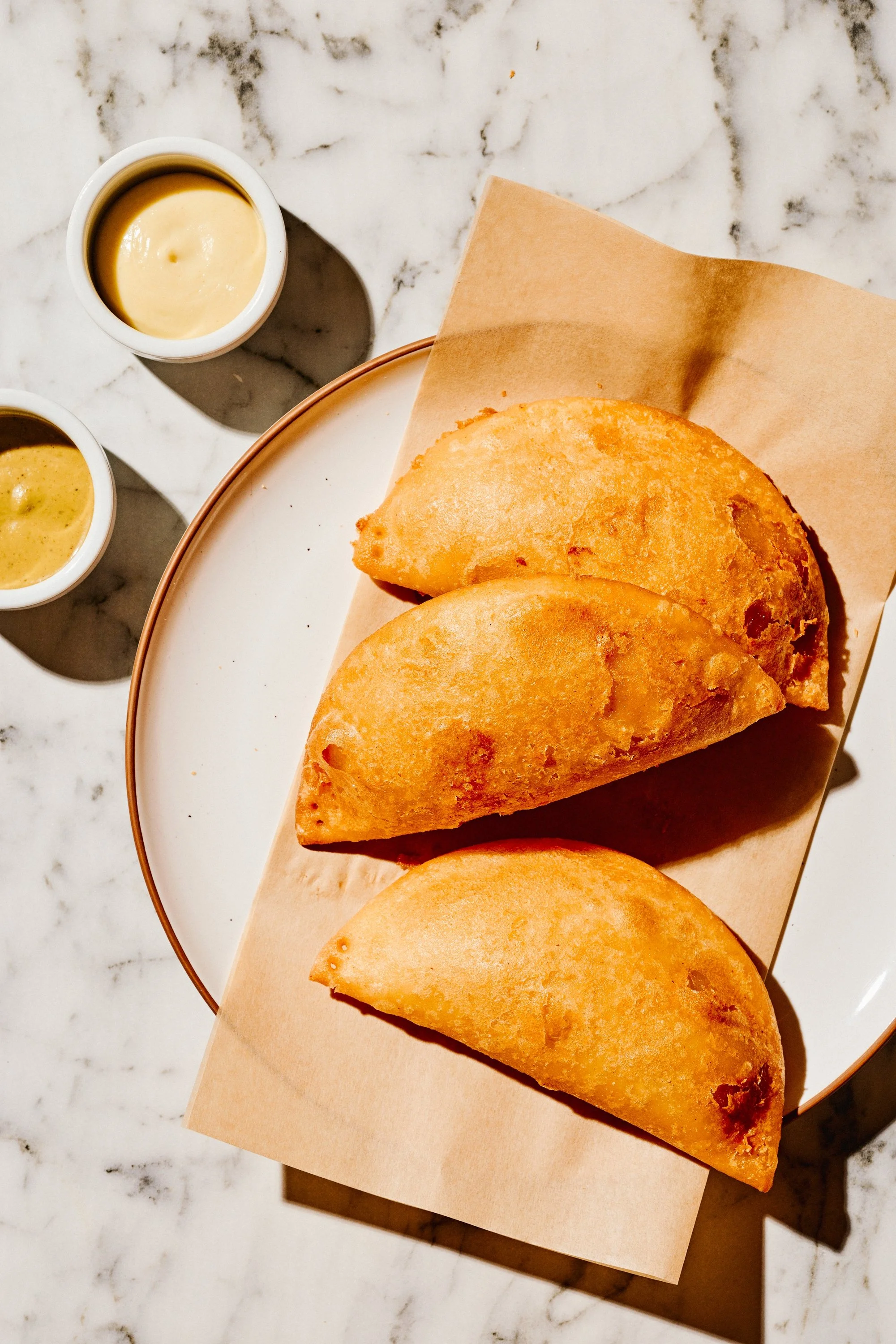 Three fried southeast Asian empanadas on a white plate with two small bowls of yellow dipping sauce, on a marble surface.