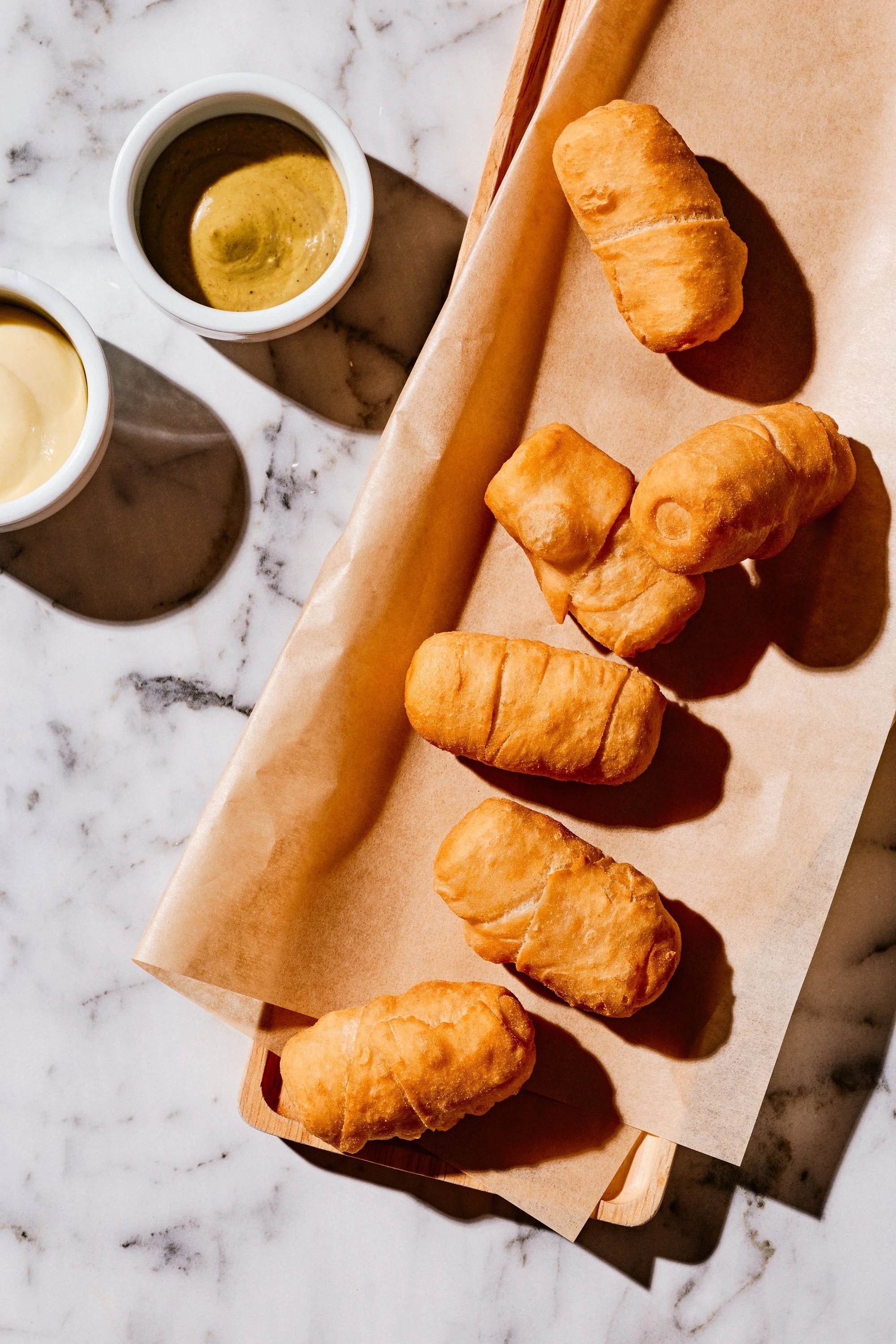 Basket of fried snacks, likely croquettes or similar, on parchment paper with two small bowls of dipping sauces on a marble surface