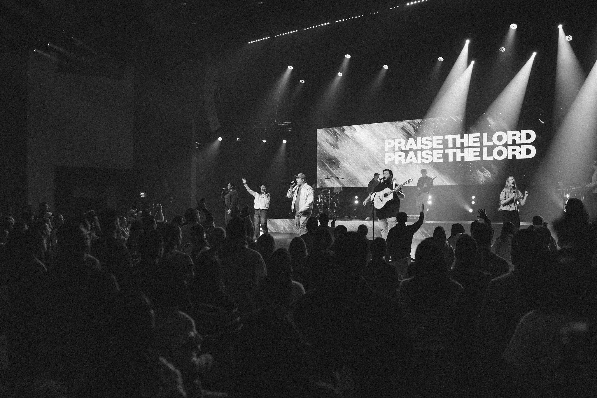 A black and white photo of a church service or concert with a group of musicians and singers on stage. The church attendees are facing the stage with some raising their hands. The background screen displays the phrase "PRAISE THE LORD" repeated twice. Several stage lights are shining down on the performers.