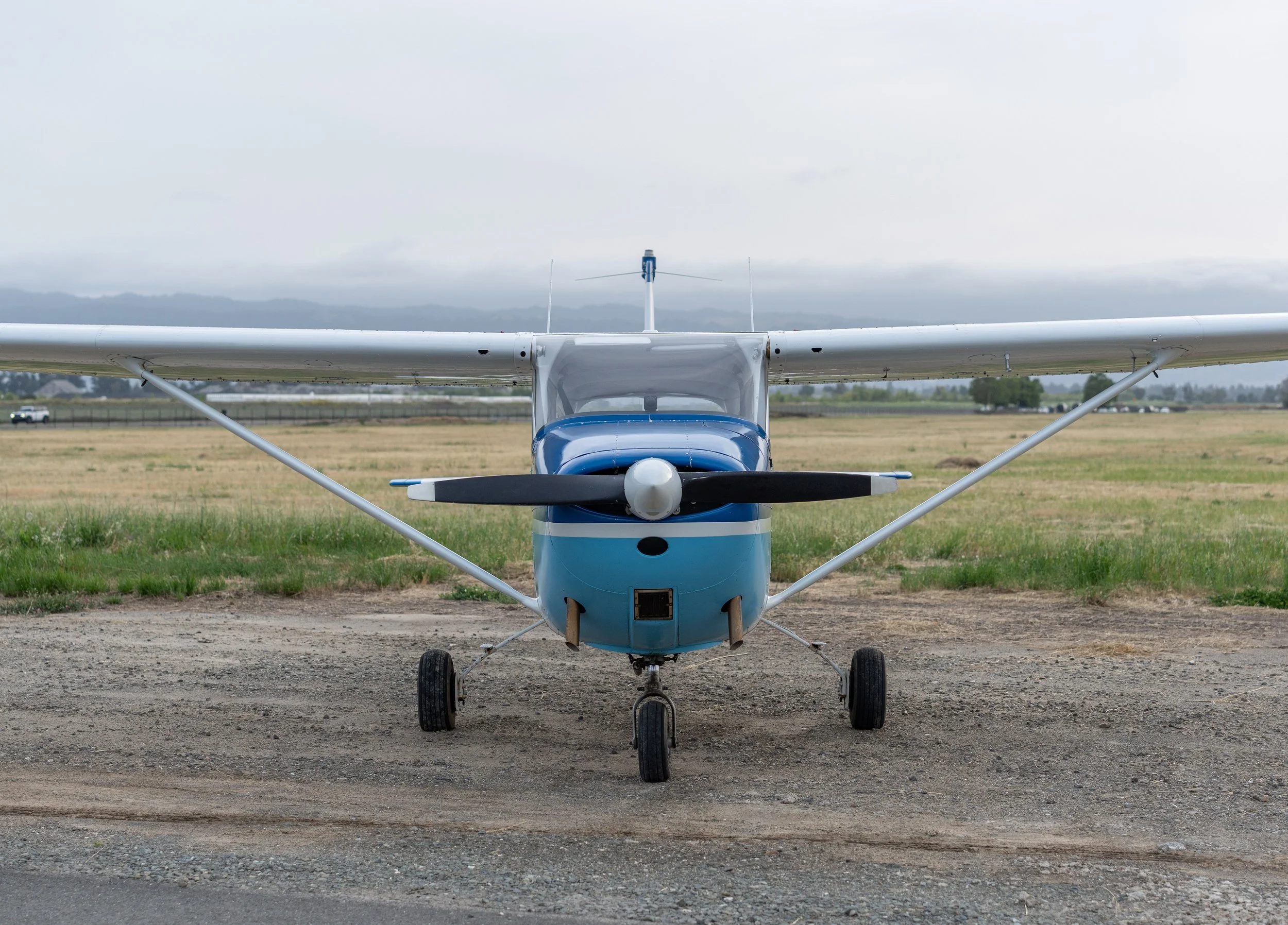 Front view of a small airplane with blue and white colors parked on a grassy airfield with mountains and cloudy sky in the background.