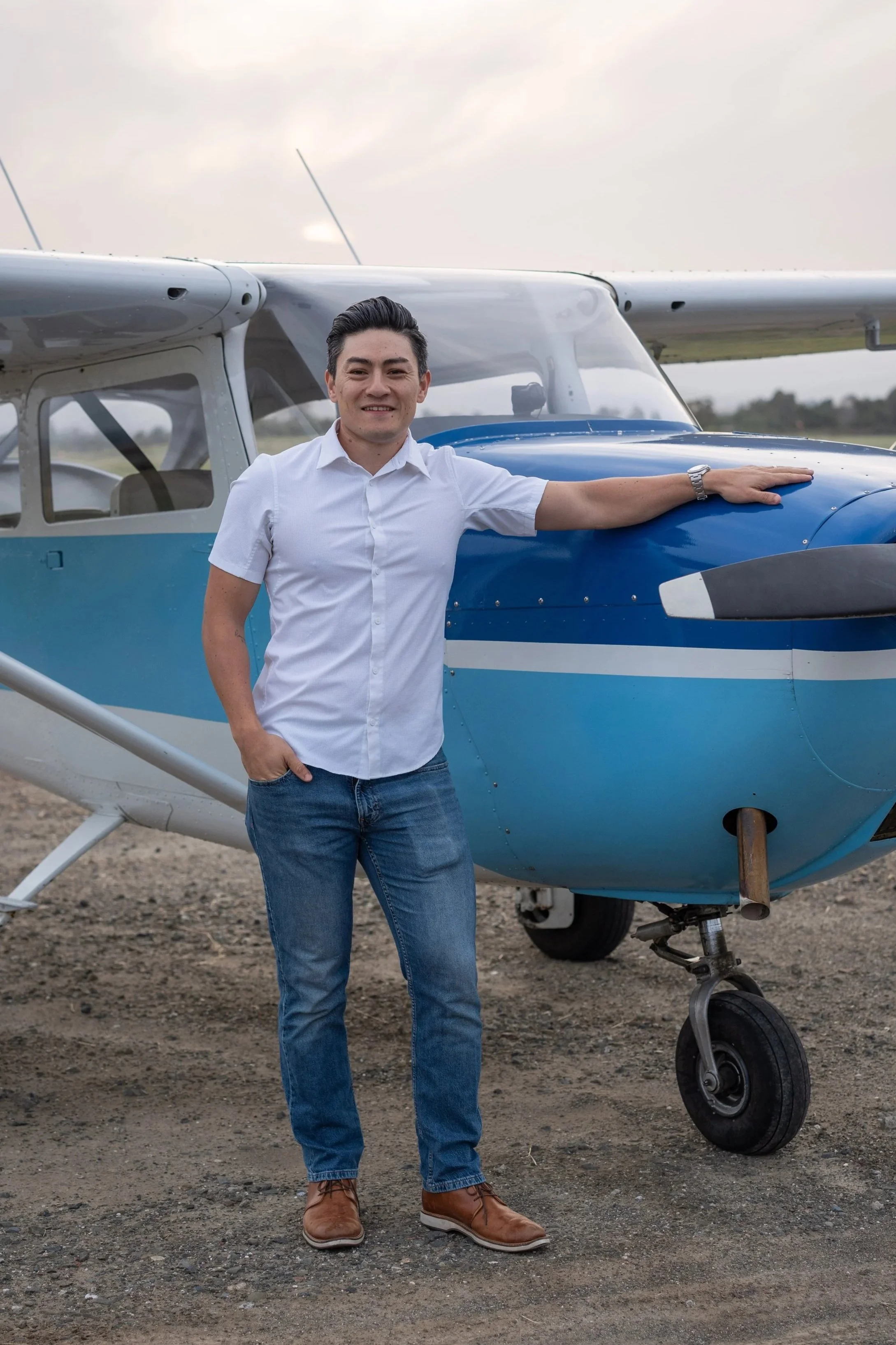 A man in a white shirt and jeans standing next to a small blue and white aircraft with its nose wheel touching the ground. He is outdoors on a dirt or gravel surface with a cloudy sky in the background.