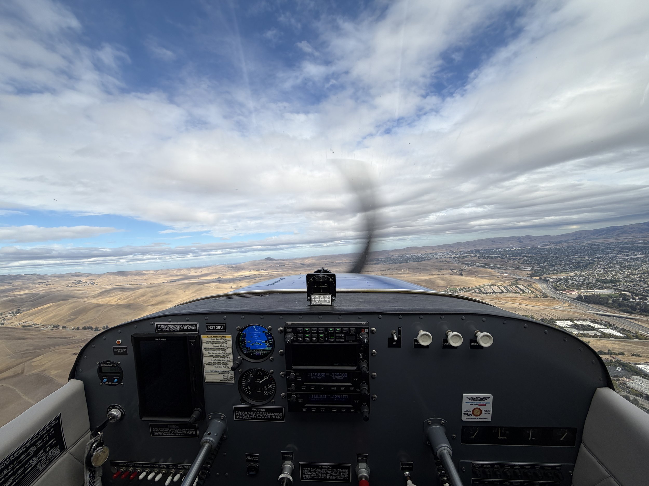 View from the cockpit of an airplane flying over a landscape with hills and a cloudy sky.