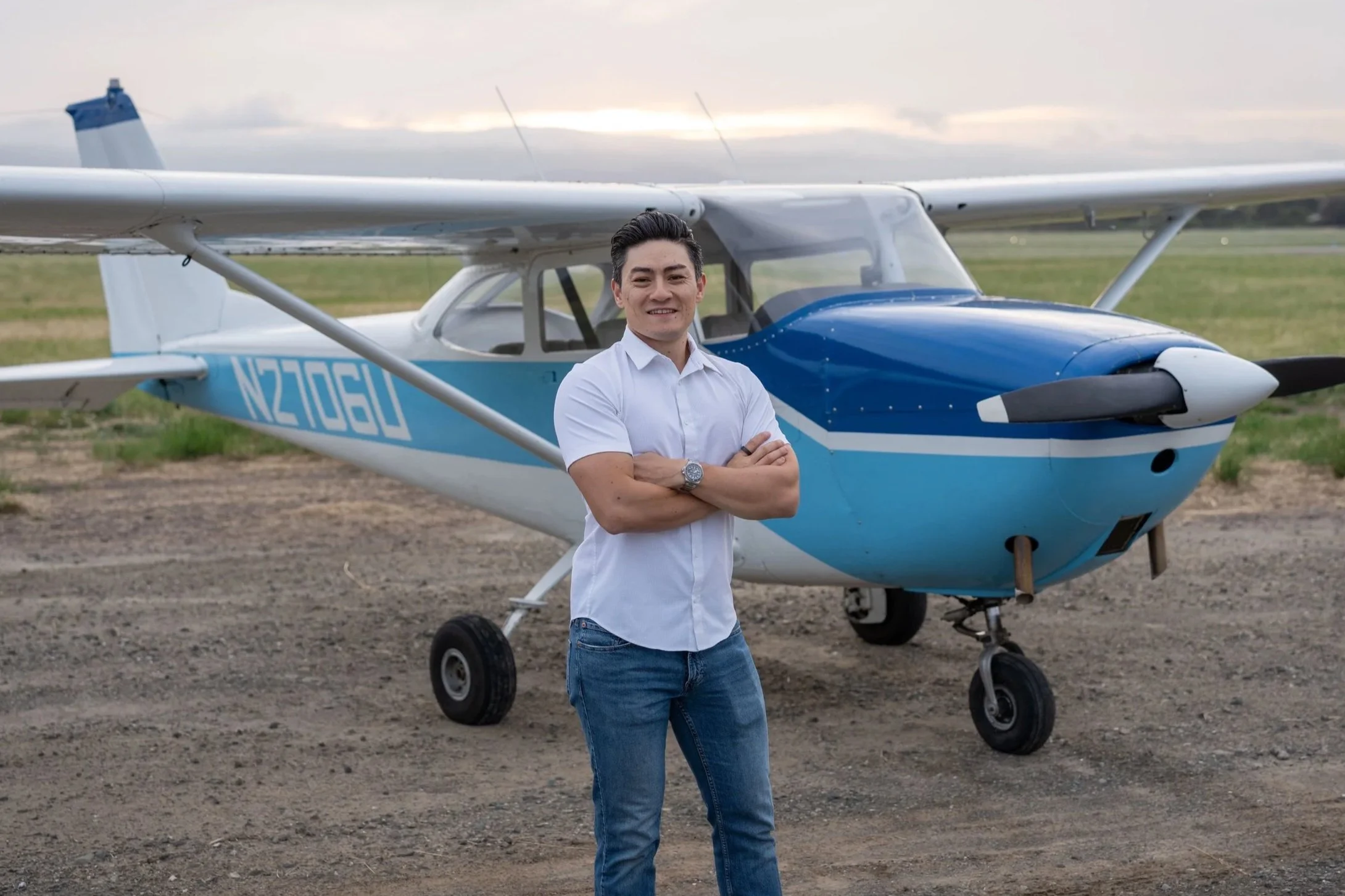 Man standing in front of a small airplane on a grassy area, with arms crossed, smiling, wearing a white shirt and jeans.