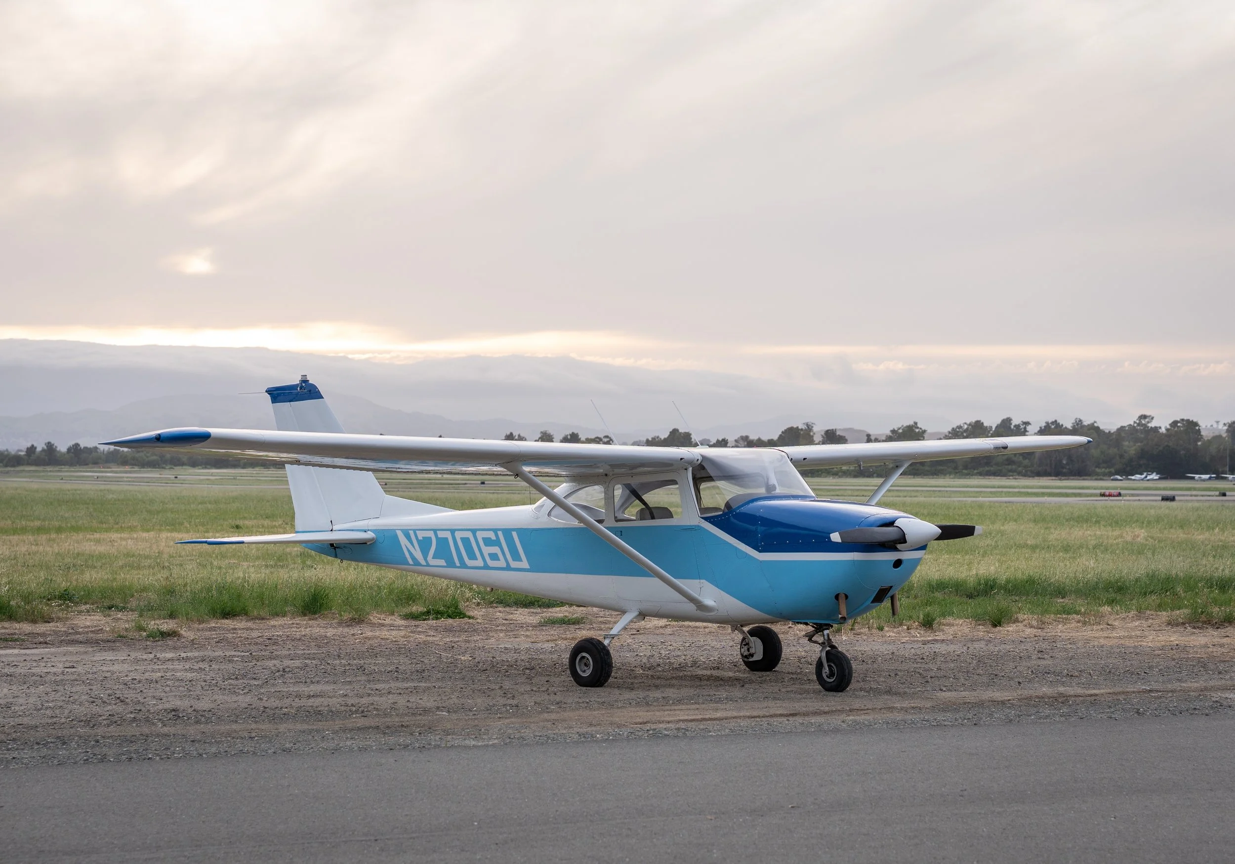 Small light blue and white aircraft parked on the grass at an airfield with overcast sky and distant mountains.