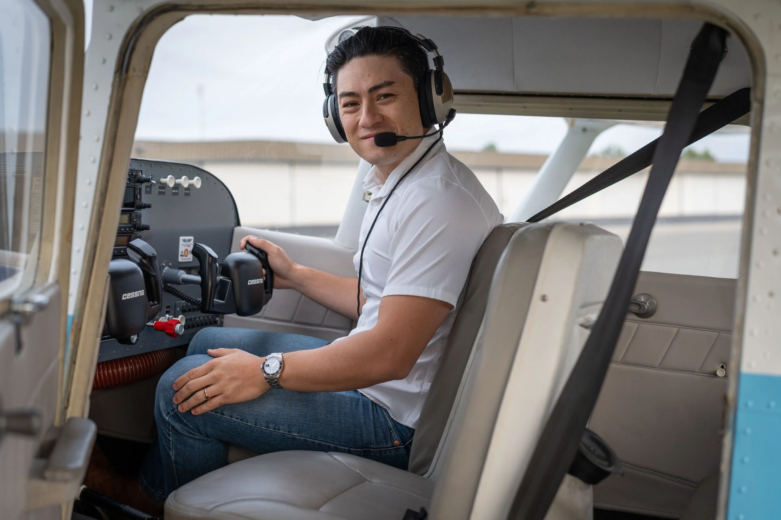 A man sitting in the cockpit of a small airplane, wearing a headset with a microphone, holding a control stick, and smiling at the camera.