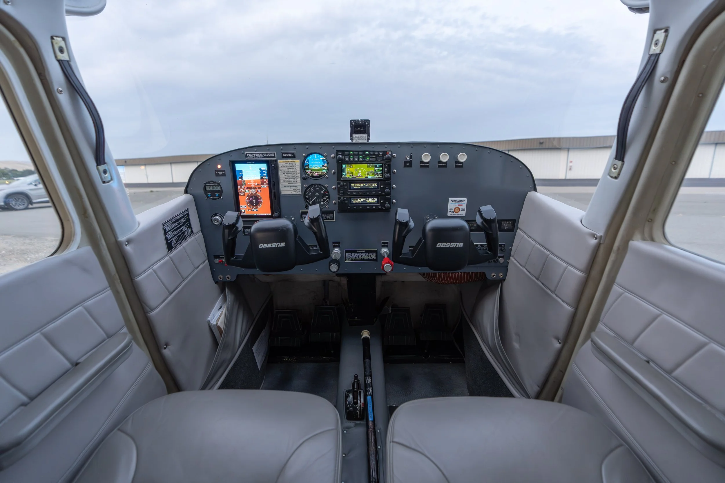 Interior of a small airplane cockpit showing various flight instruments, displays, and controls, with a view of the airport tarmac and sky through the front window.