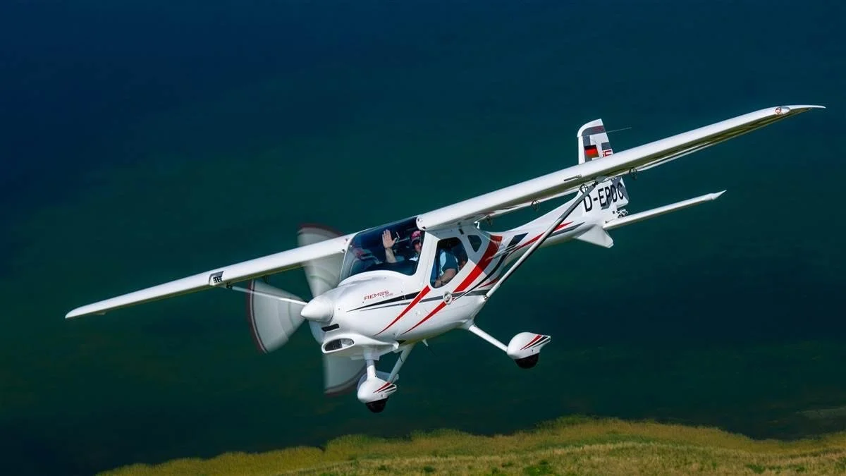 Small white aircraft flying low over grassy terrain with a pilot waving from the cockpit.
