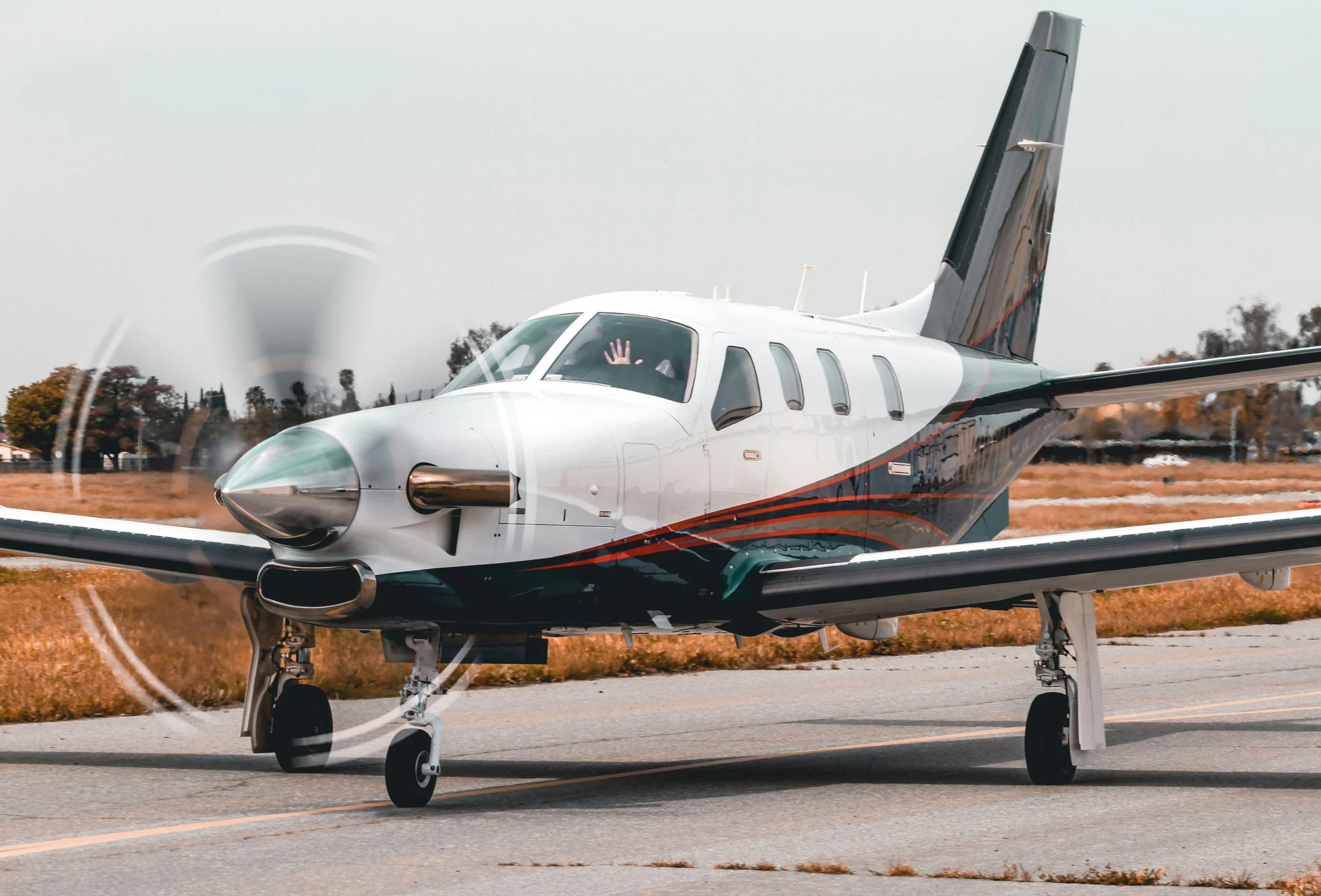 A small private jet aircraft on a runway, with a pilot waving through the cockpit window. Trees and clear sky in the background.