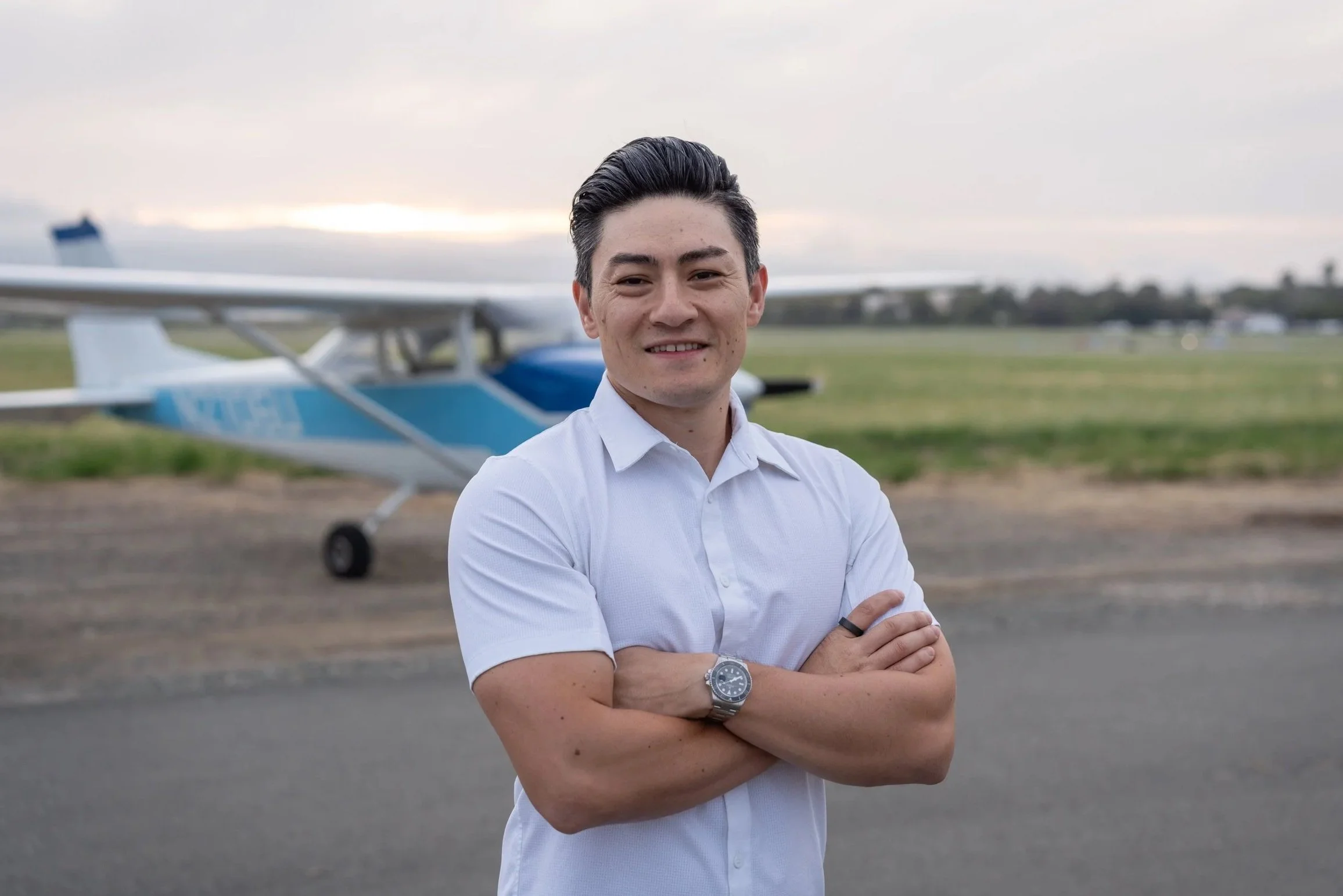 A young man with dark hair and light skin, wearing a white shirt and a watch on his left wrist, standing outdoors in front of a blue and white airplane on a grassy airfield during overcast weather.