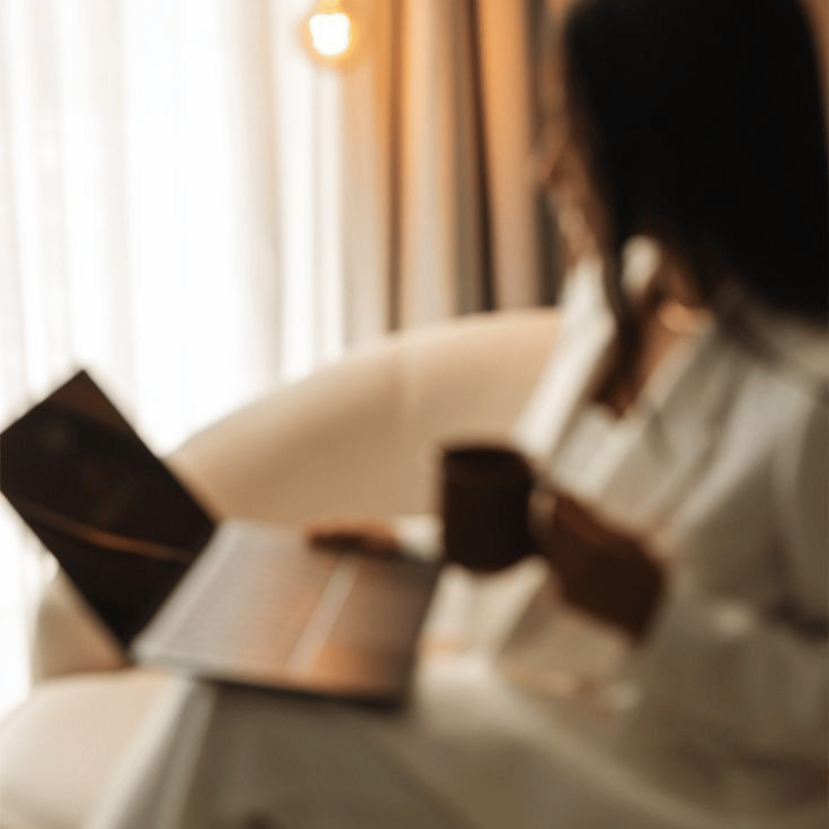 A woman sitting on a cream-colored couch reading a book and drinking coffee, with a tablet or e-reader nearby, in a cozy room with curtains and warm lighting.