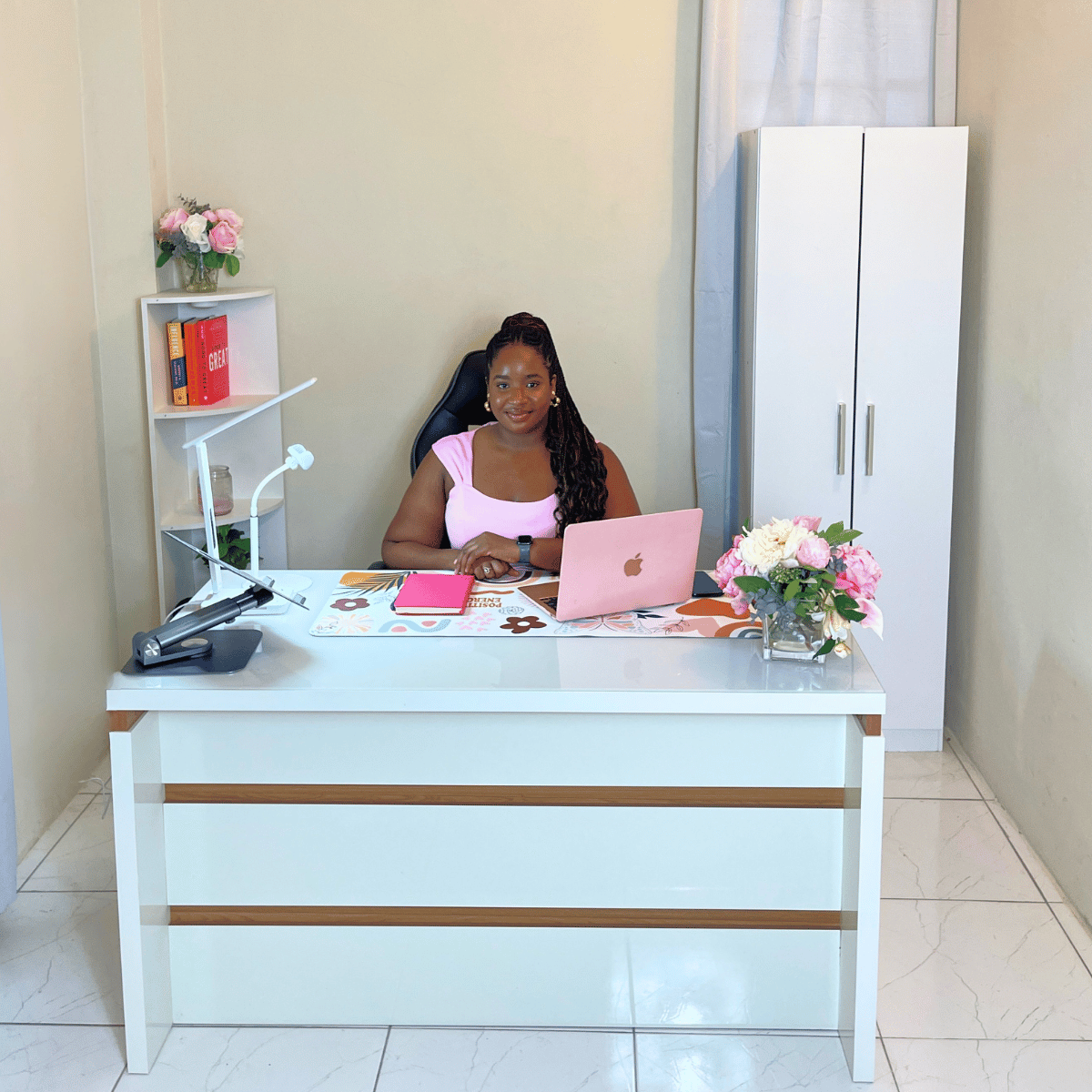 A woman sitting at a white desk in an office, with a pink laptop, pink notebook, a desk lamp, and a vase of pink and white flowers.