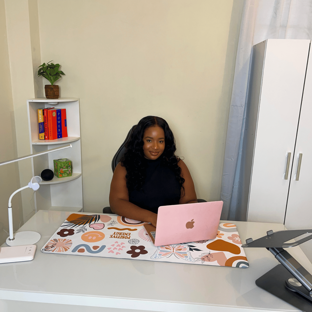 Woman with long black curly hair sitting at a desk with a pink MacBook, in a room with a white wardrobe, a white shelf with books and decor, and a white desk lamp.