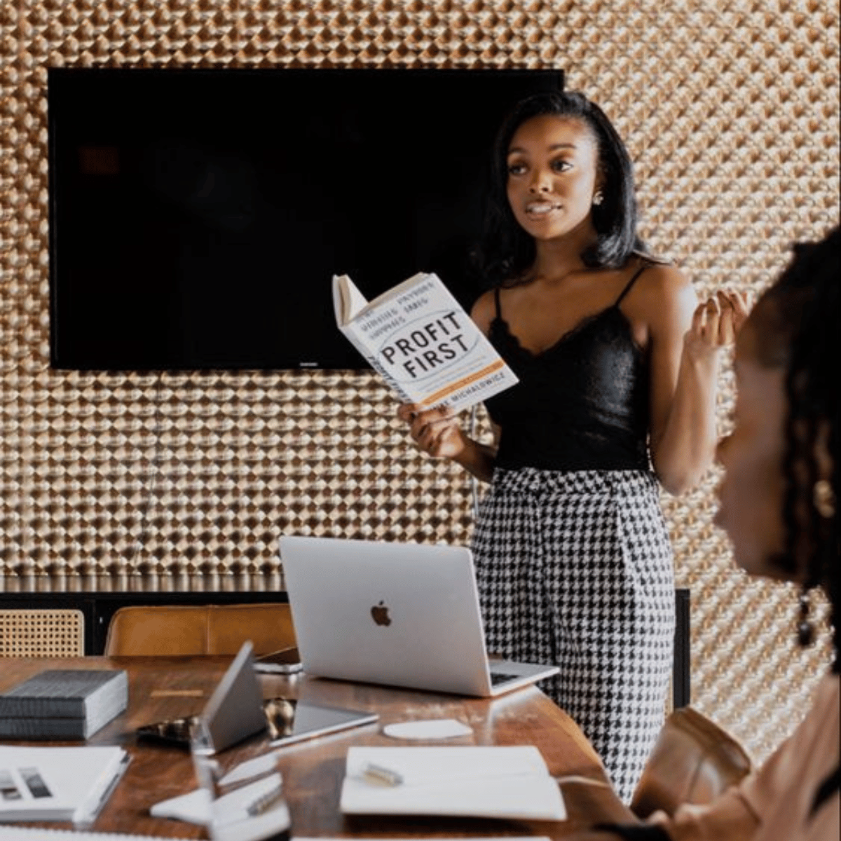 A woman in a black top and checkered pants is giving a presentation in a conference room. She is holding a book titled "Profit First" and gesturing with her right hand. There are laptops and papers on the table.