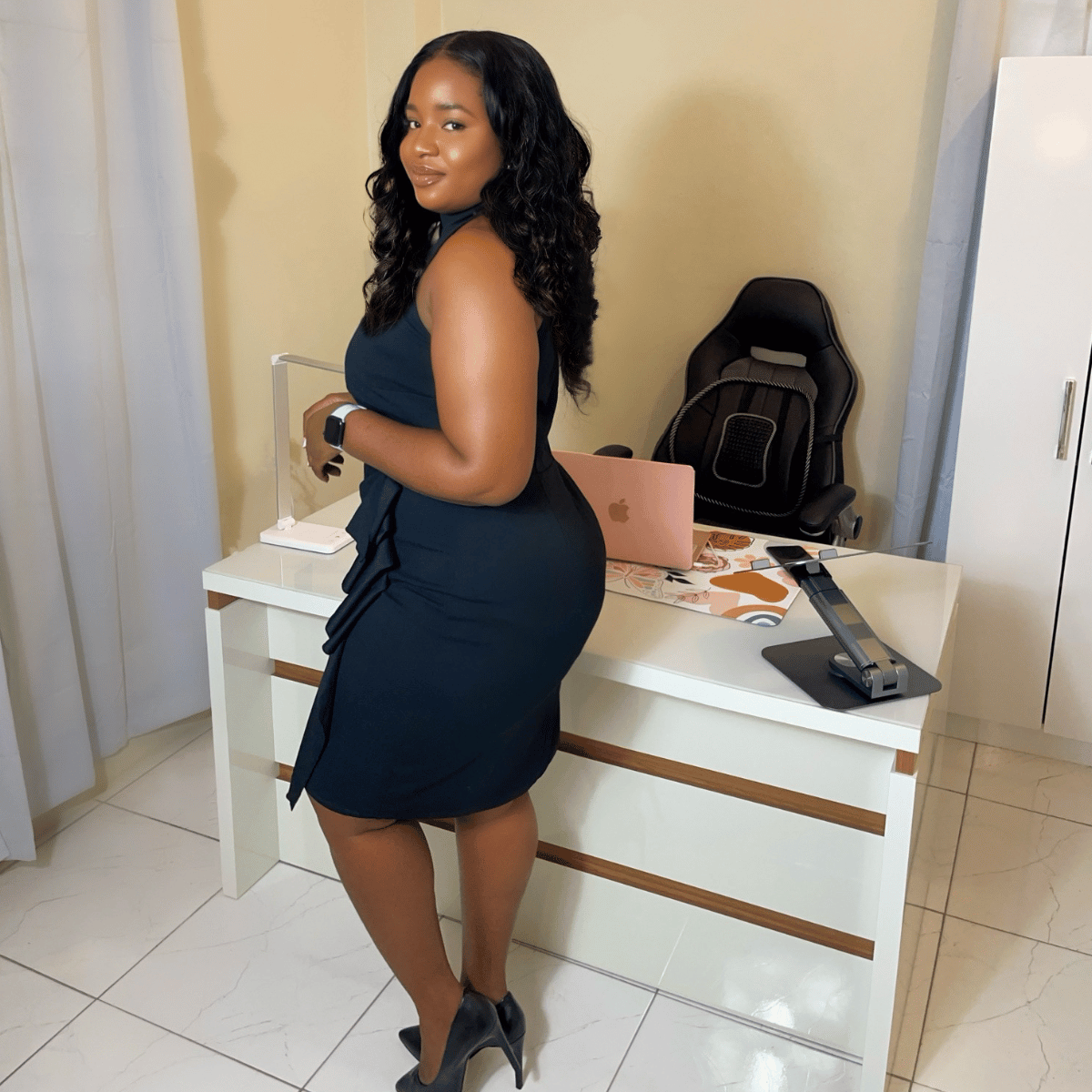 A woman with long dark hair wearing a black dress and high heels standing in a room with a desk, laptop, and chair.