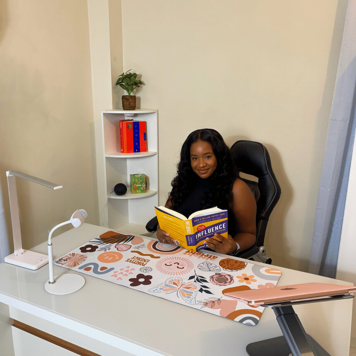 Woman sitting at a desk reading a book titled 'Influence' in an office setting with a white desk, colorful floral desk mat, a small white shelf with books and decorative items, and a beige wall.