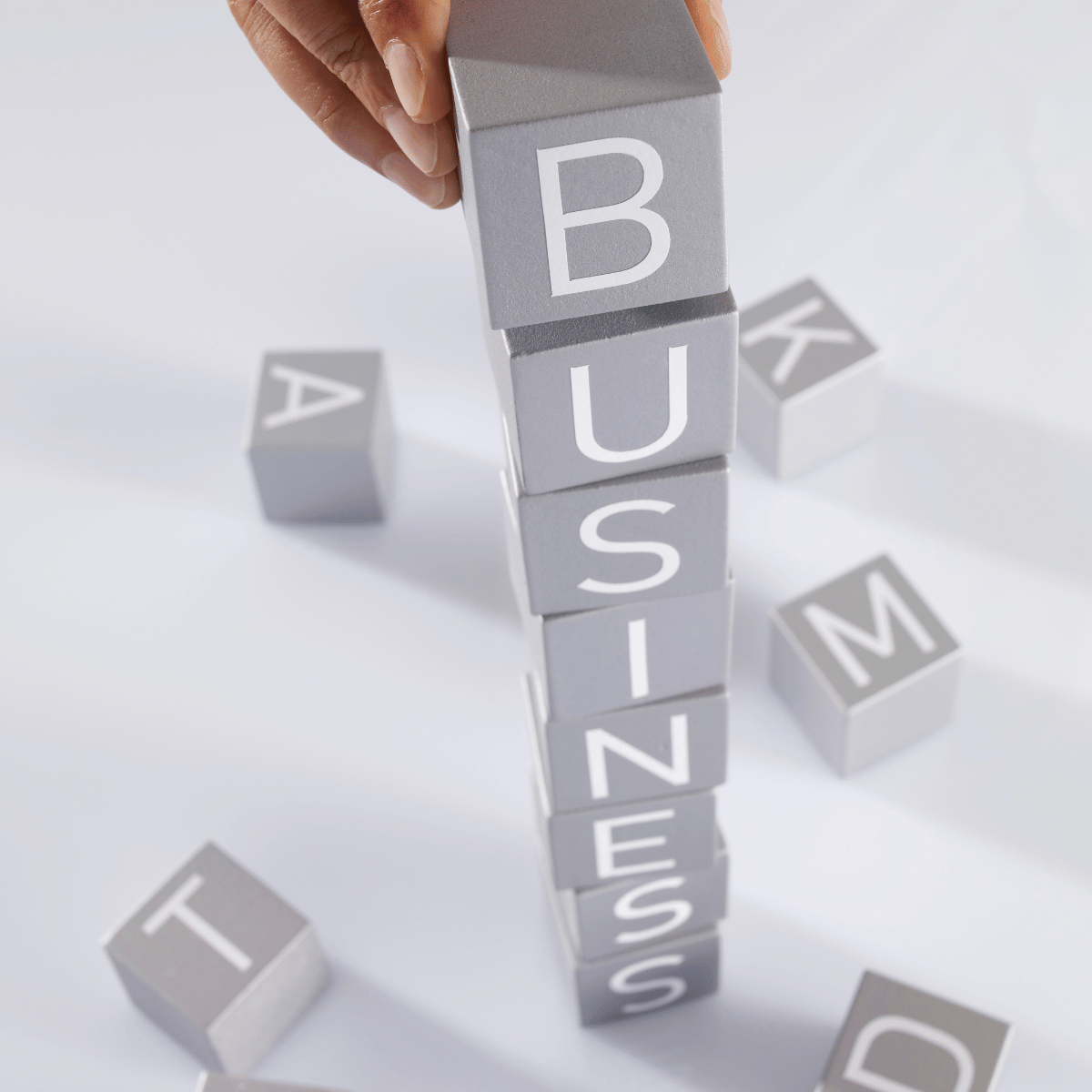 Hand stacking gray blocks with white letters spelling 'BUSINESS', with scattered blocks with letters on a white surface.