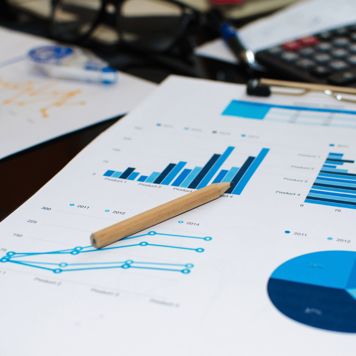 Close-up of printed business charts and graphs on paper, with a wooden pencil, a calculator, and eyeglasses on a desk.