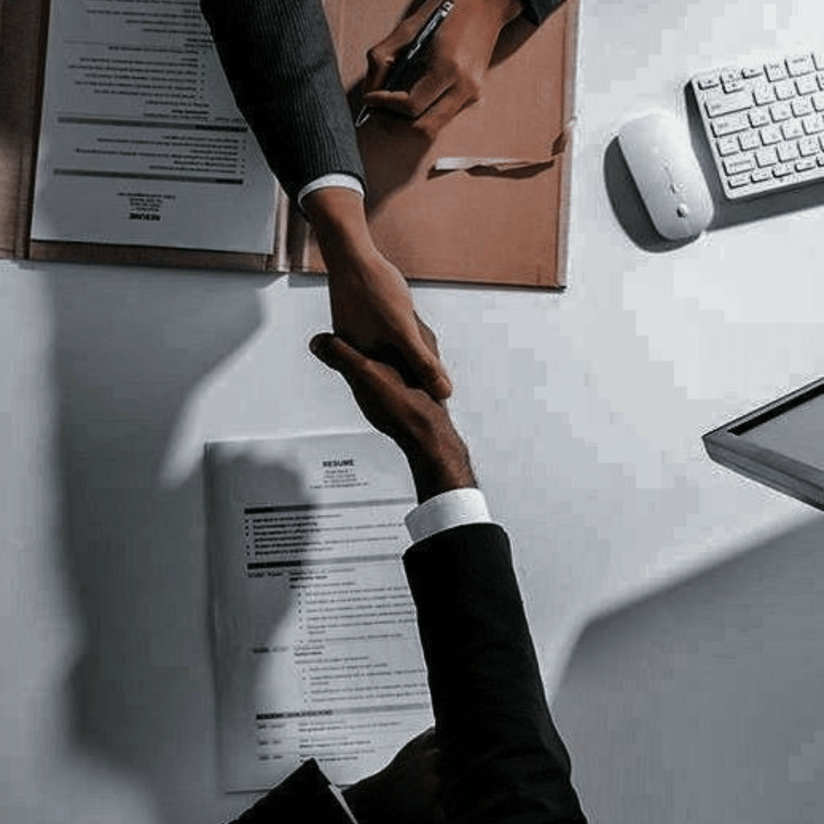 Two people shaking hands over a desk with documents, a computer keyboard, and a mouse, in an office setting.