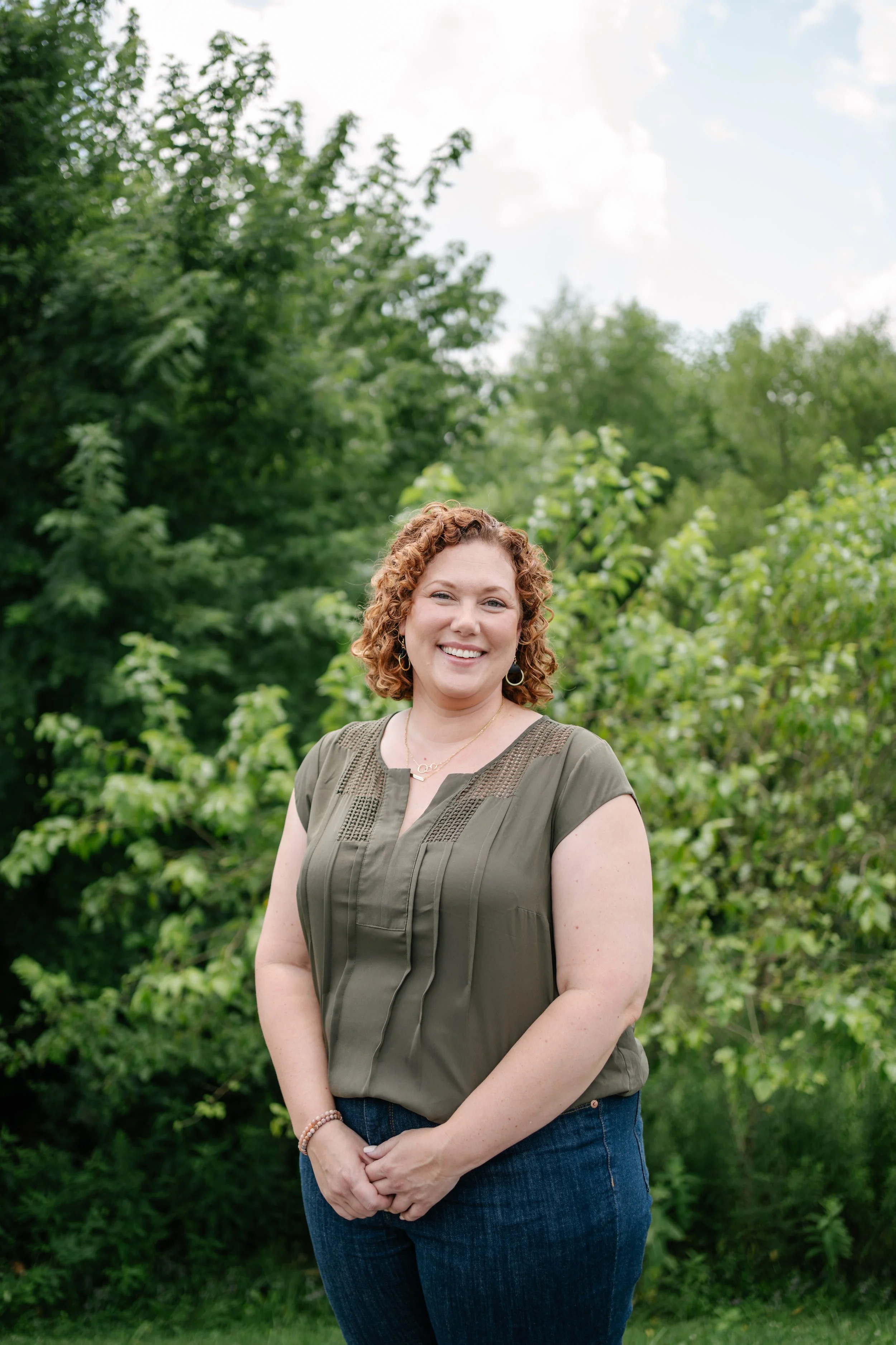A woman with curly red hair standing outdoors in front of green trees, smiling and wearing a green blouse and jeans.