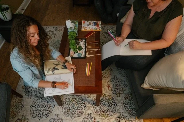 A woman and a girl are sitting at a small wooden table, each with sketchbooks and colored pencils, engaging in drawing or coloring activities.