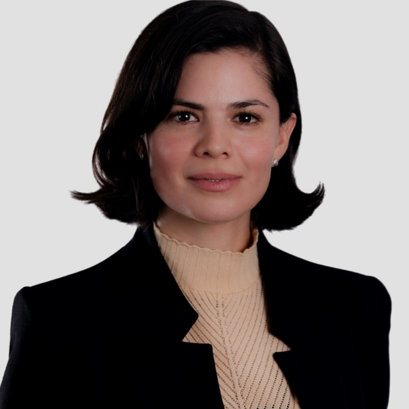 Mujer con cabello corto, vestida con blazer negro y suéter beige, mirando hacia la cámara en fondo gris.